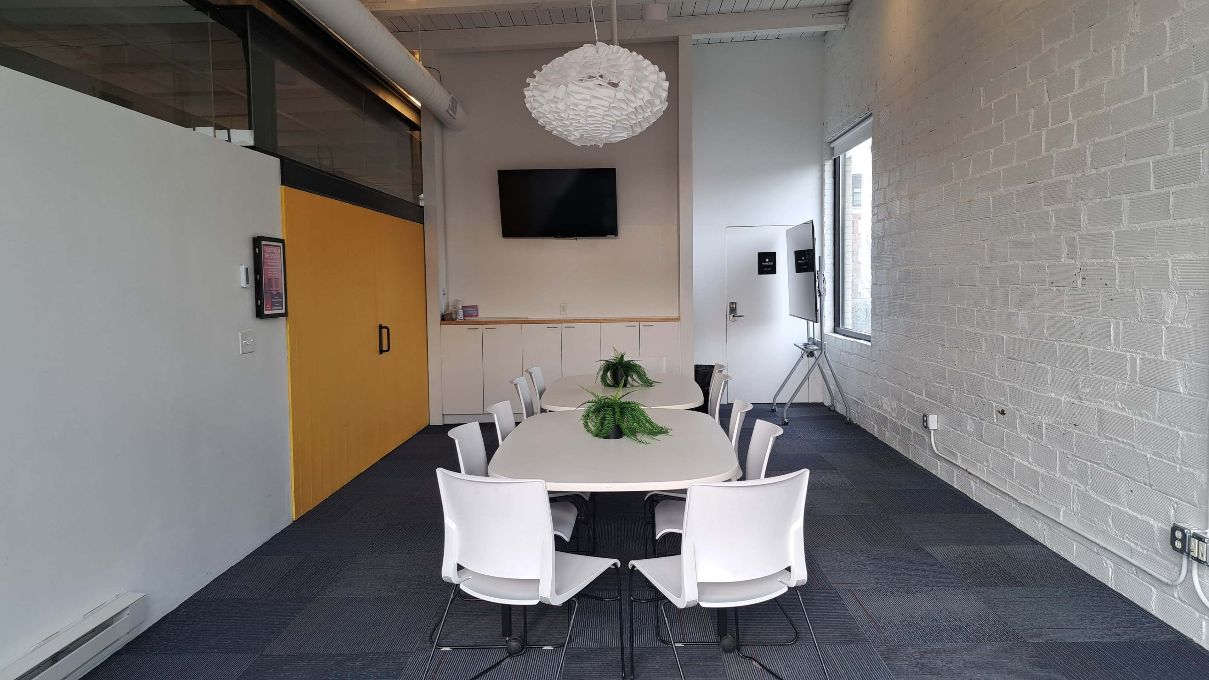 A conference room features a long white table surrounded by eight chairs, with a large pendant light overhead and a television mounted on the wall.