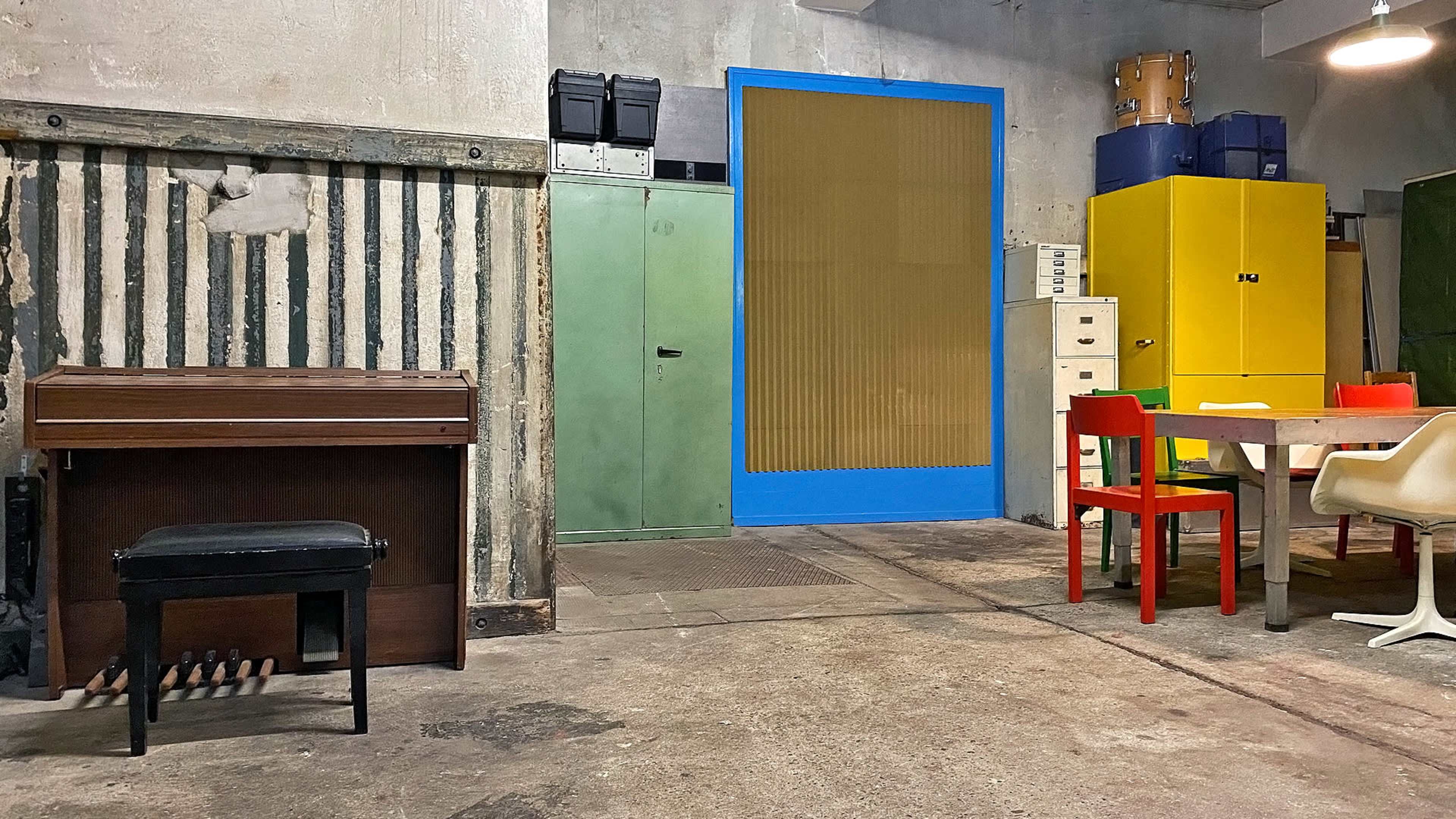 A room features a wooden piano, colorful cabinets, a table with chairs, and a cement floor.