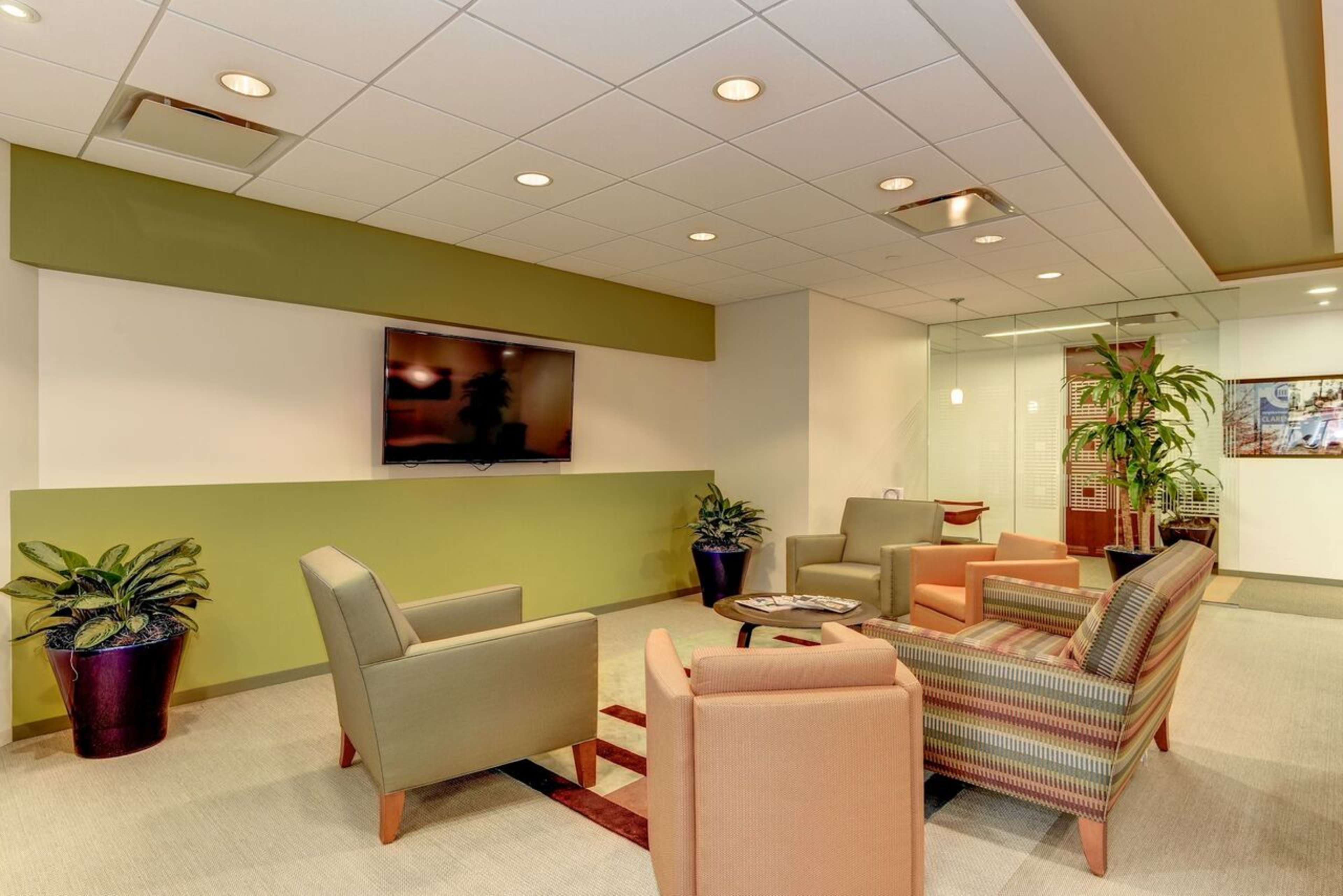 A modern office lounge area furnished with multiple chairs, a coffee table, and a television mounted on a green-accented wall, alongside potted plants.