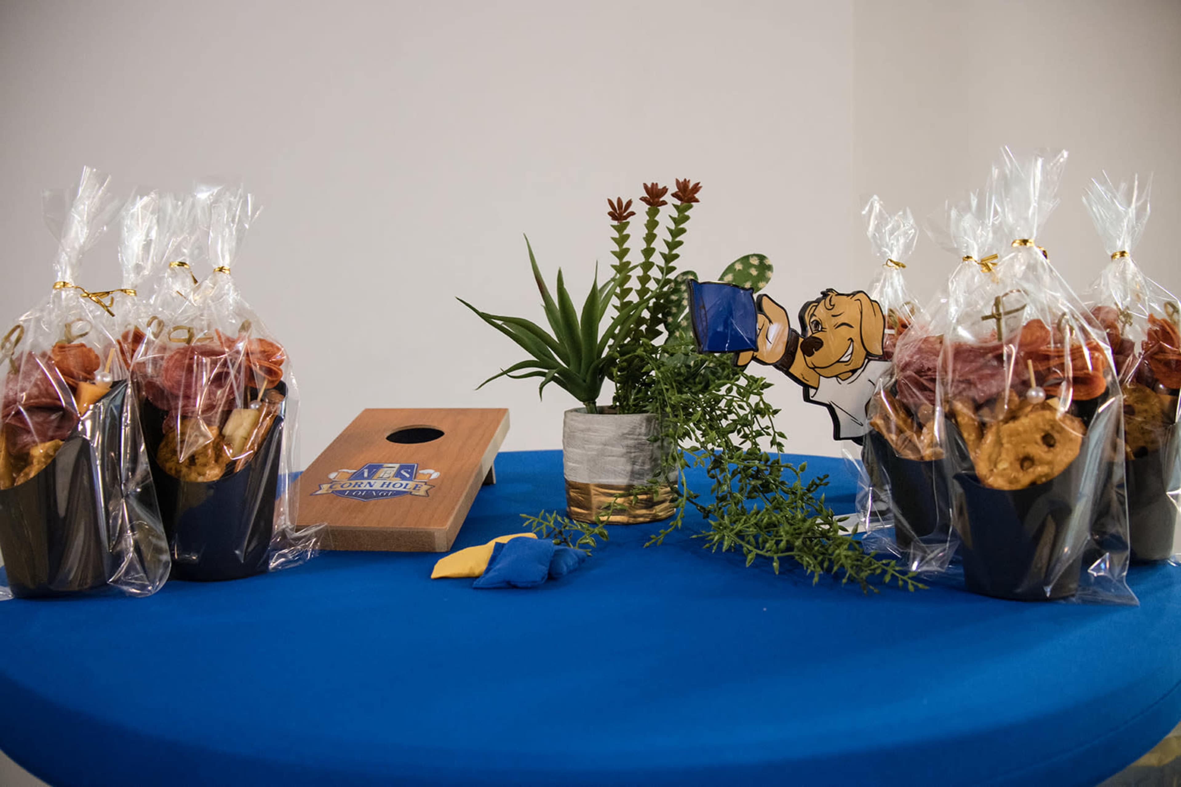 A blue table displays several gift bags filled with snacks, a small potted plant, and a wooden box with a cutout figure pouring from a watering can.