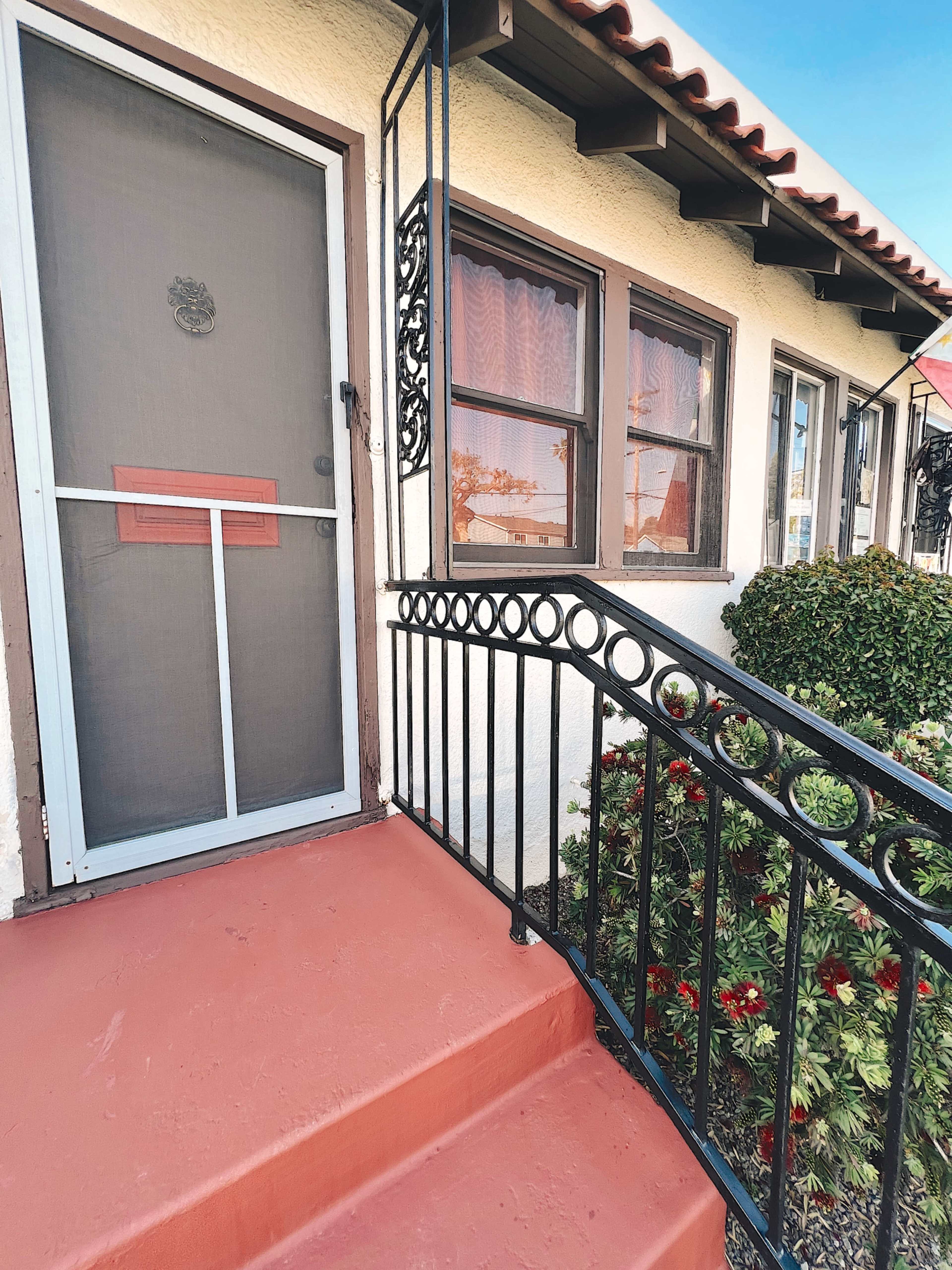 The image shows a front entrance with a screened door, a decorative railing, and a red-painted step leading to a small garden area.