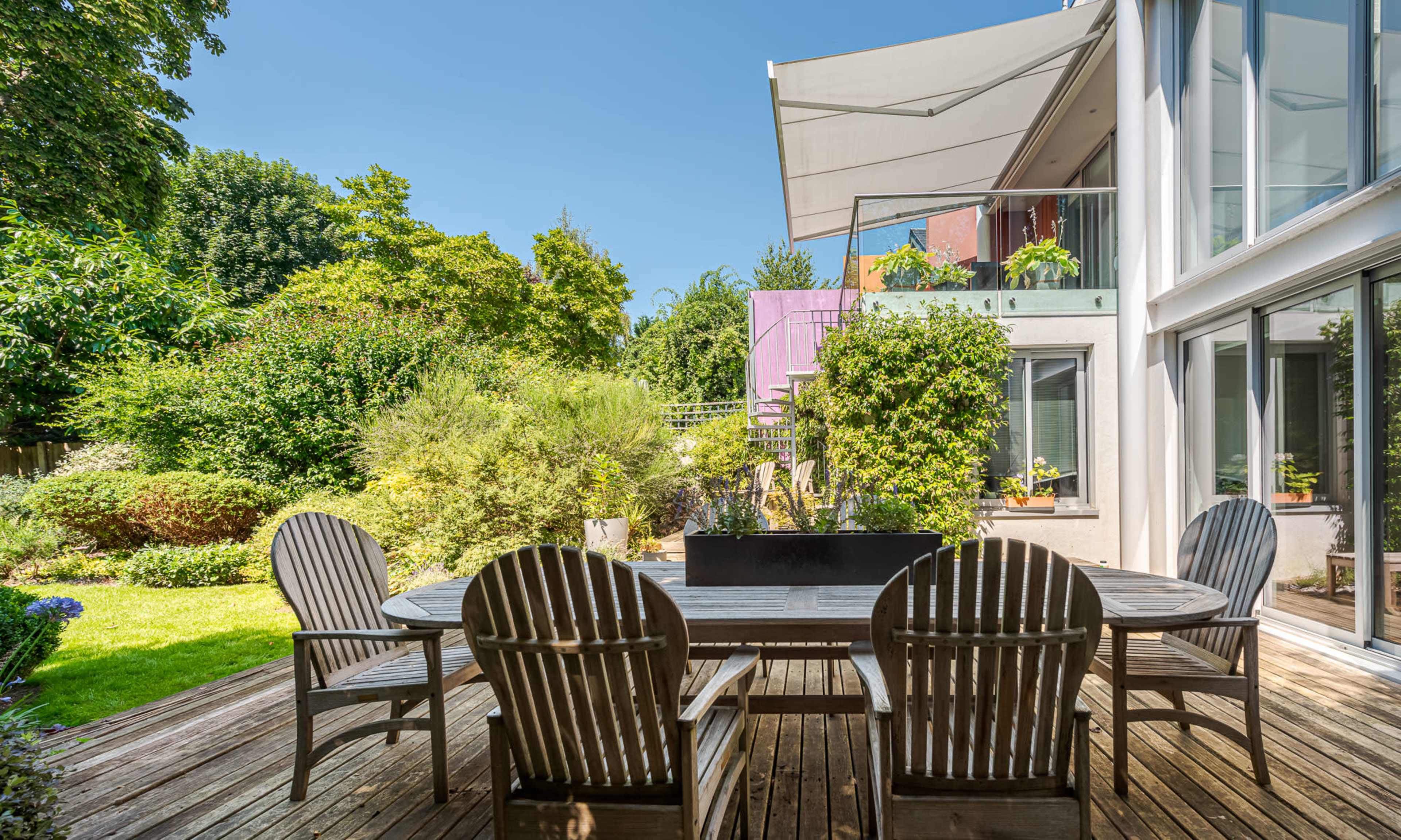 The image shows a wooden patio with a dining table and chairs, surrounded by a lush garden and a modern house featuring large windows.