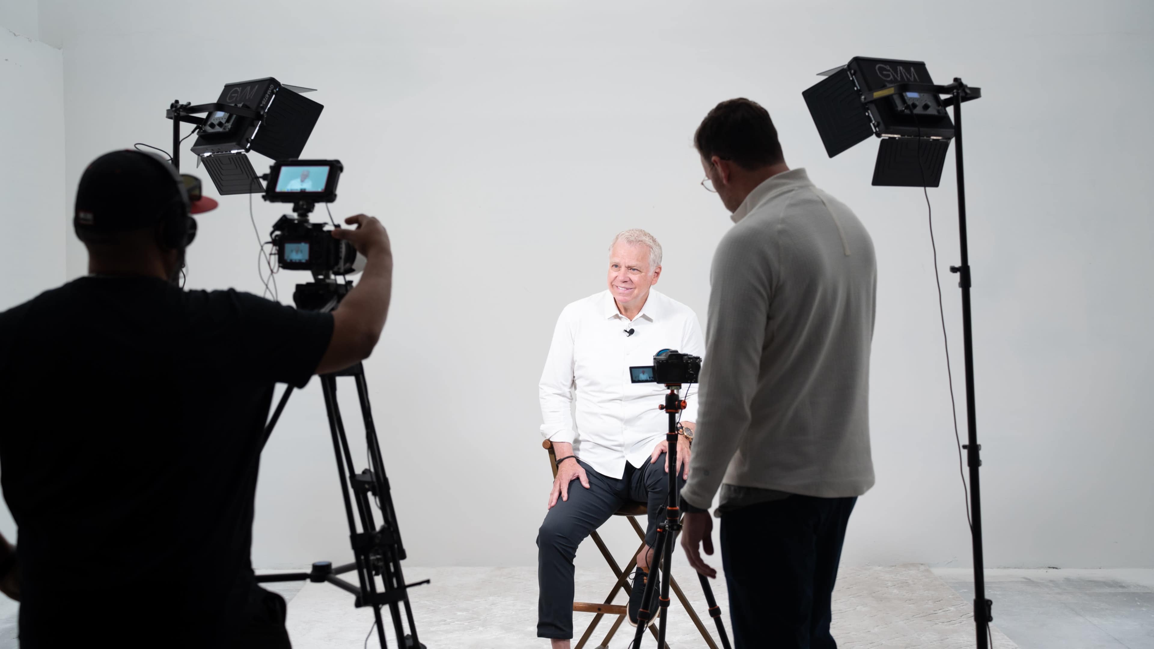A man sits on a stool in a studio setting while being filmed by a crew using multiple cameras and lights.