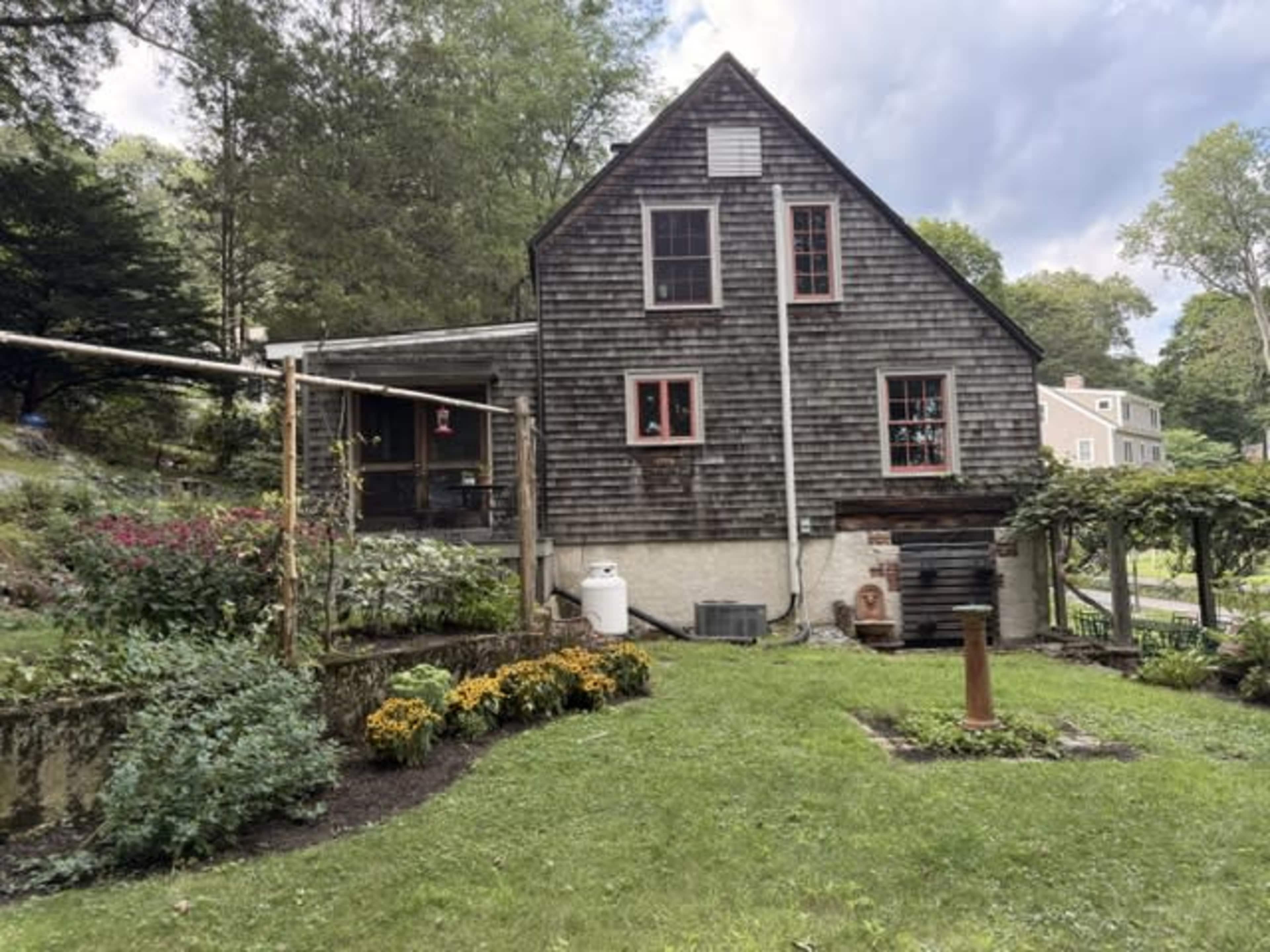 The image shows a two-story, wooden house with a dark shingled exterior and red trim, surrounded by a grassy yard and landscaped flower beds.