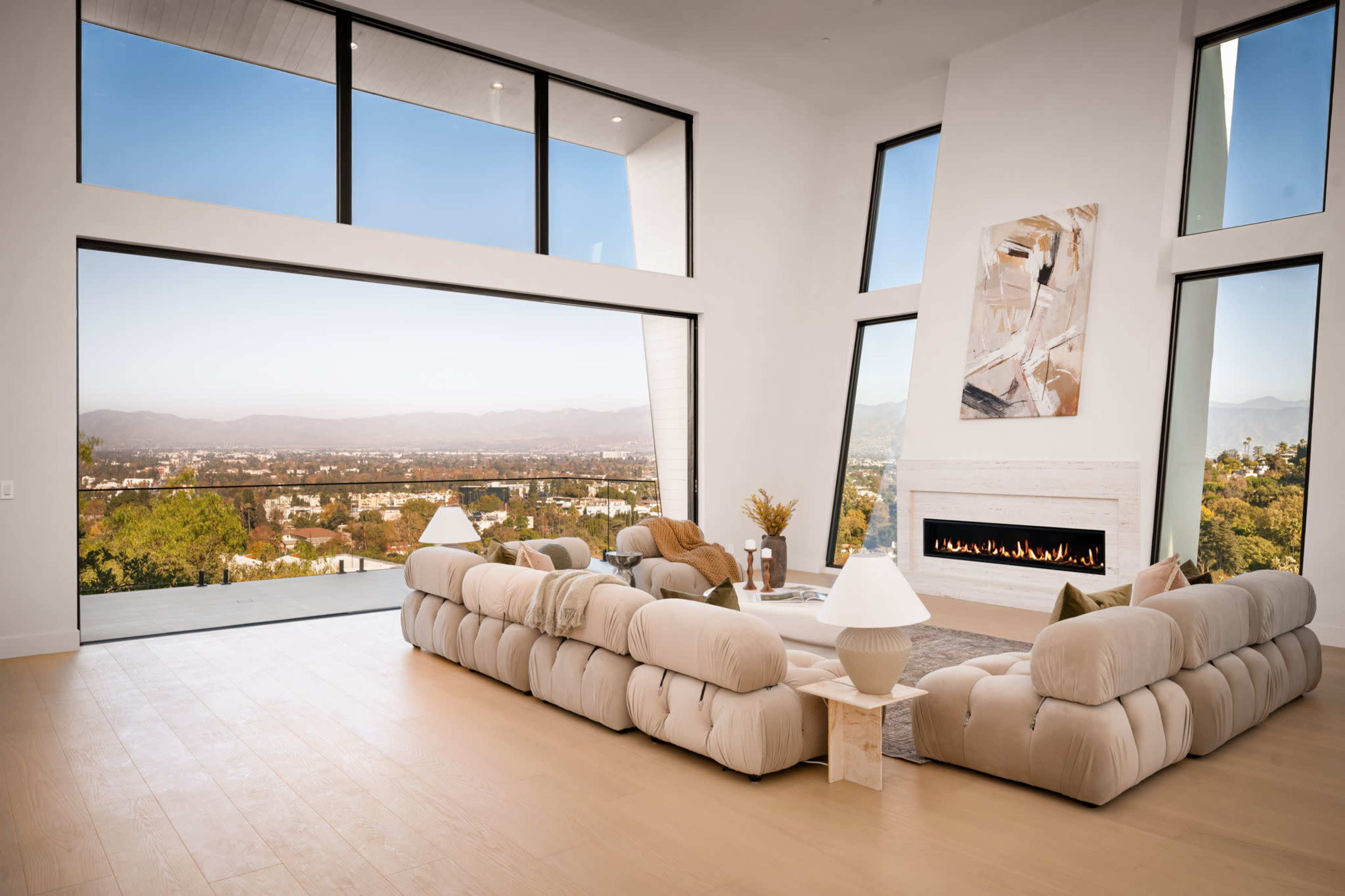 The image depicts a modern, spacious living room with large windows showcasing a scenic view of hills and a cityscape, featuring a beige sectional sofa and a minimalist fireplace.