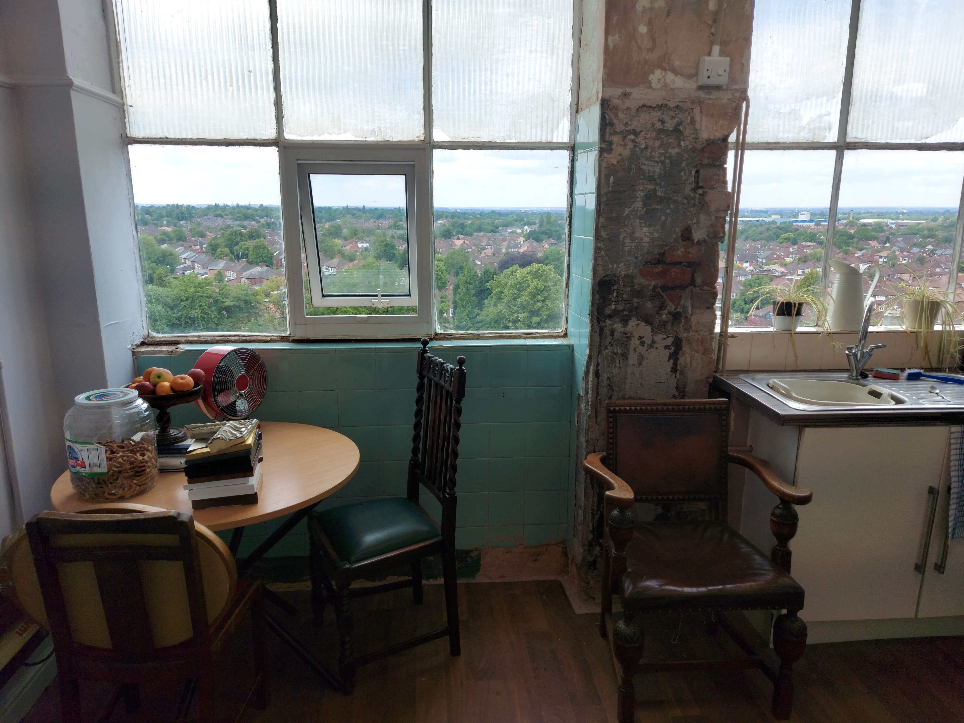 A small room features a table with fruit and a fan, two chairs, a kitchen area, and large windows providing a view of the surrounding landscape.