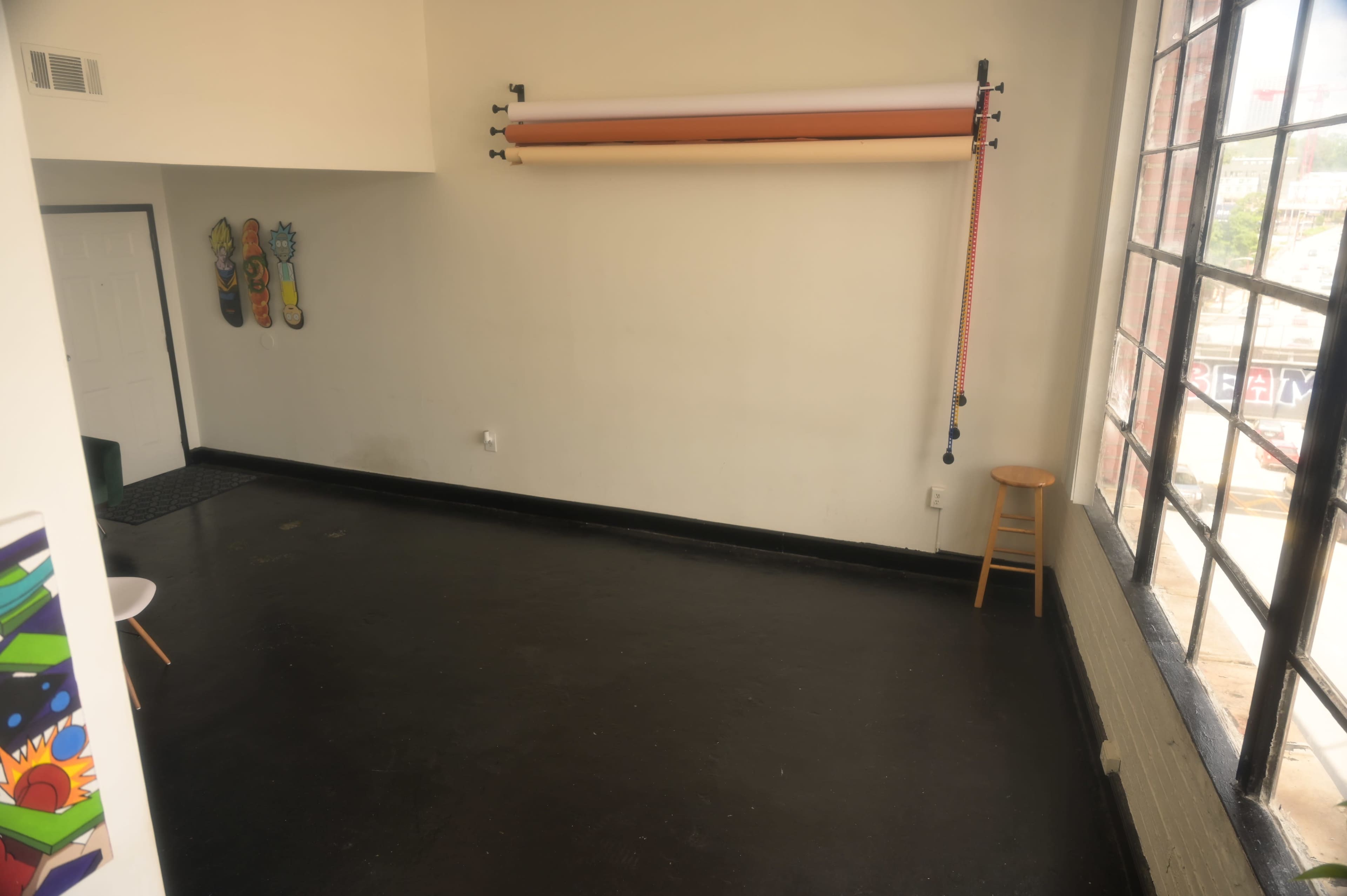 A spacious, empty room with a black floor, a stool in the corner, and colorful artwork on the wall.
