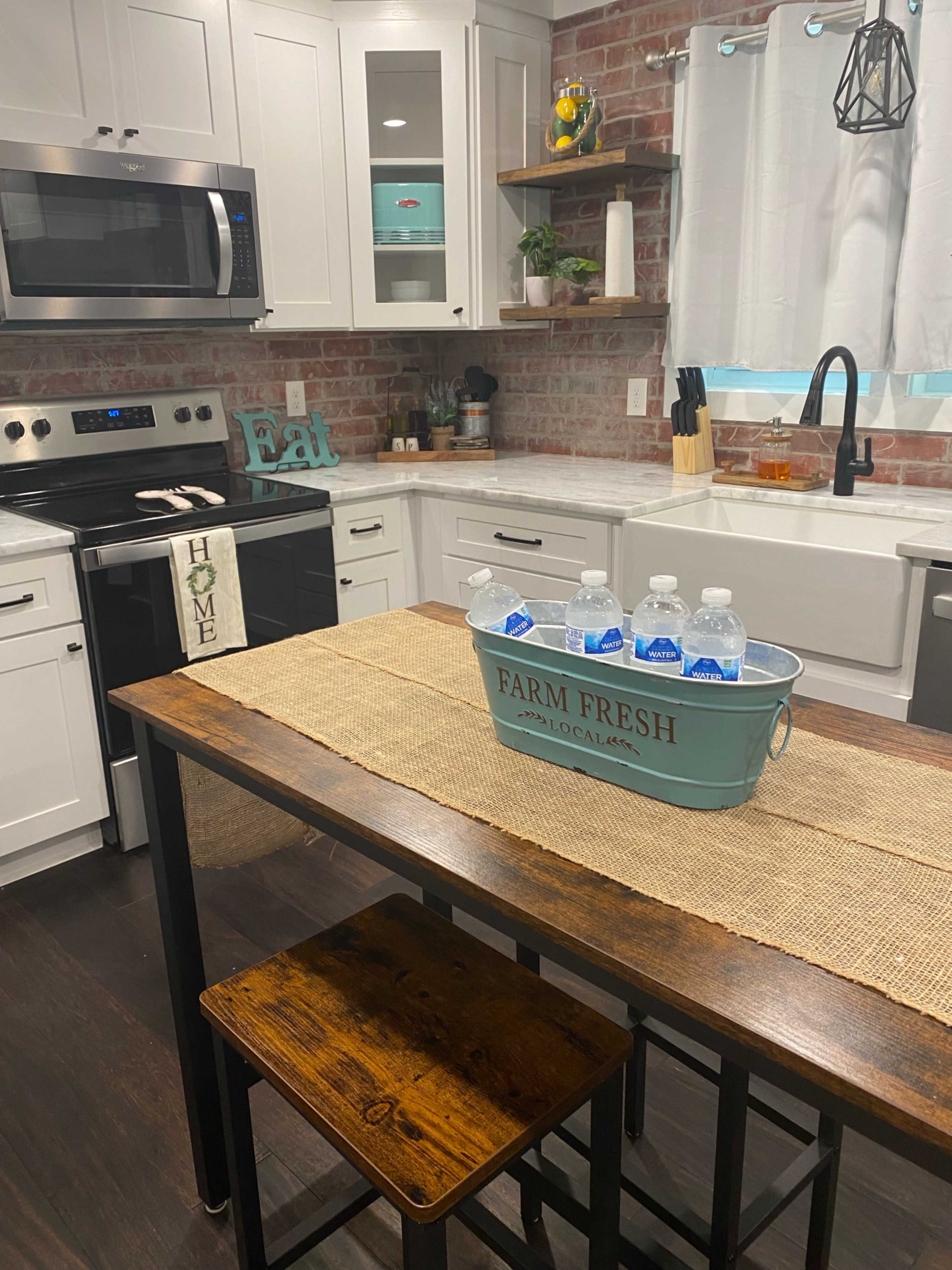 The image shows a modern kitchen featuring white cabinetry, a brick accent wall, a wooden dining table with a blue metal basket holding water bottles, and a black sink.