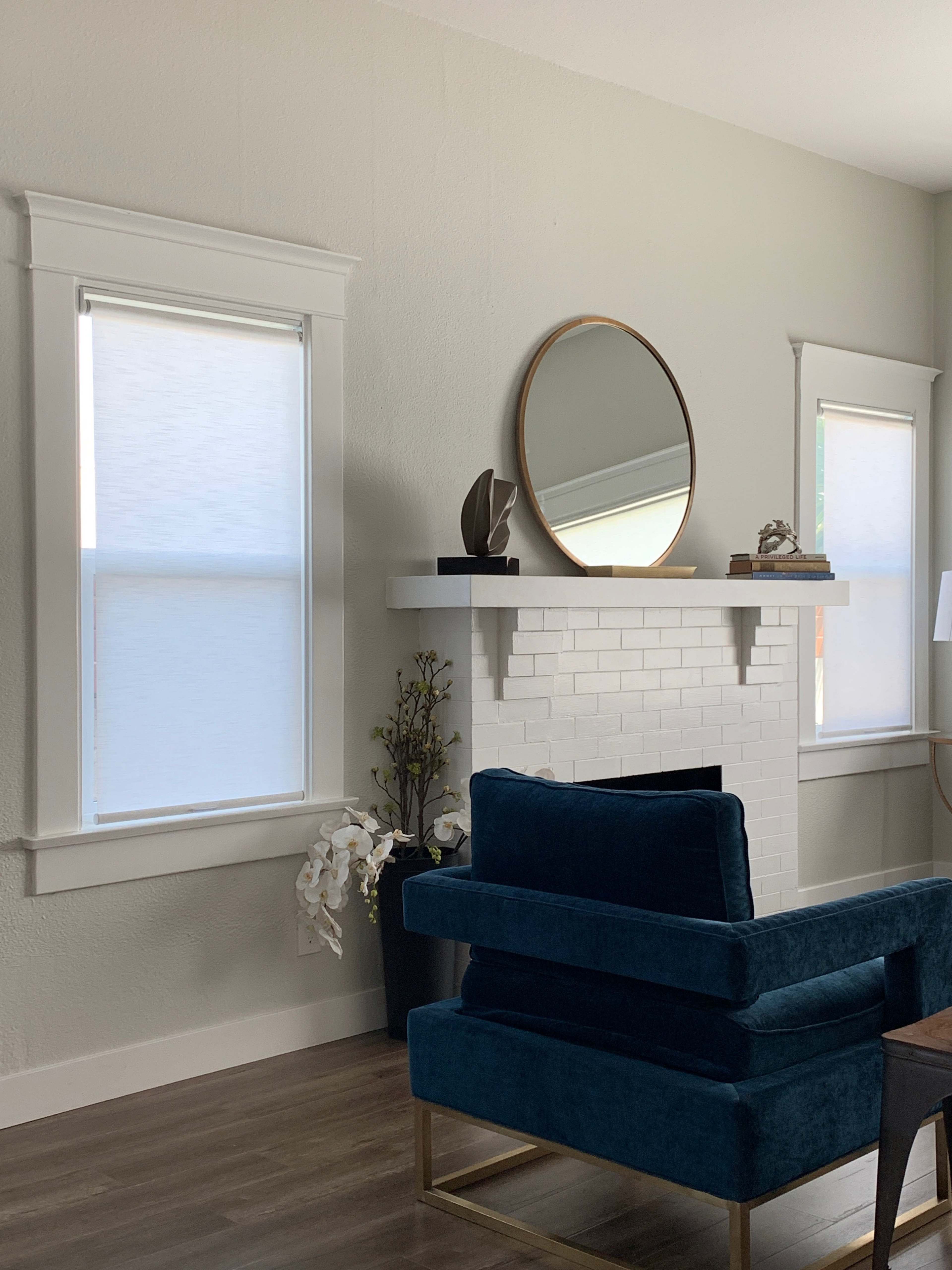 The image shows a cozy living room featuring a white tiled fireplace with a round mirror above it, a blue velvet chair, and window shades on either side.