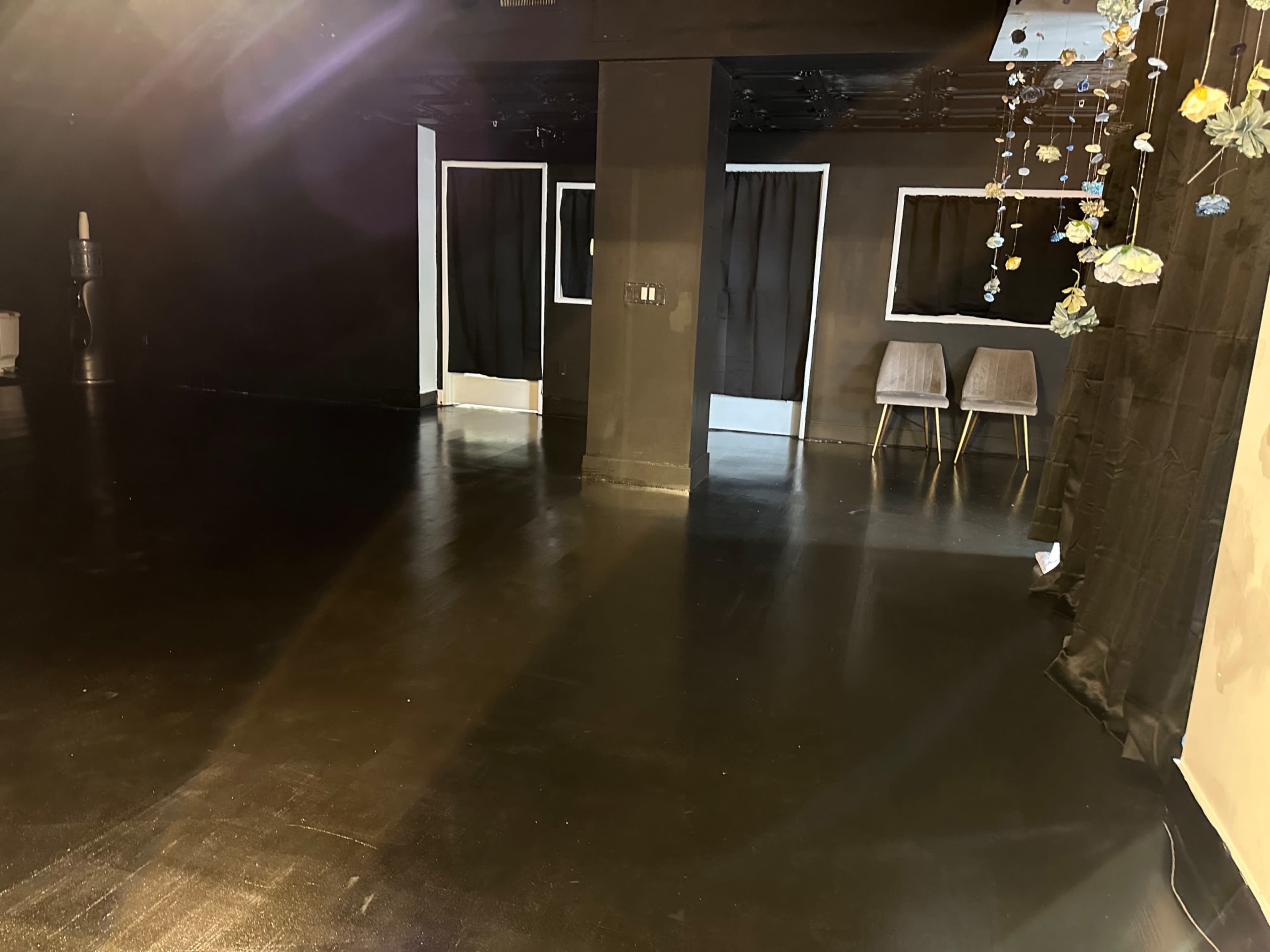 A dimly lit room with a black floor, black curtains, and two chairs placed against a wall.