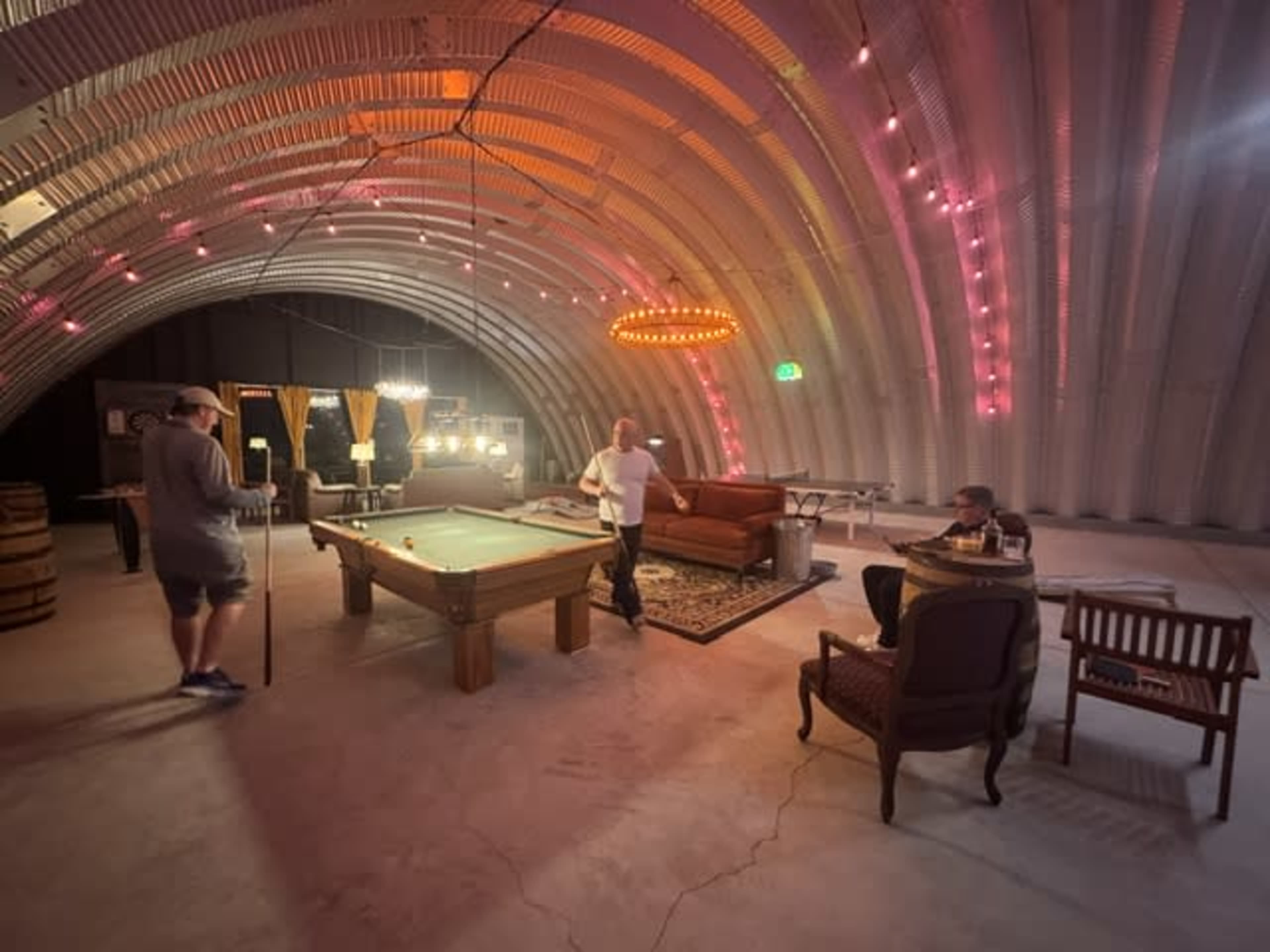 The image shows a spacious interior with a pool table, seating areas, and ambient lighting in a curved metal building.