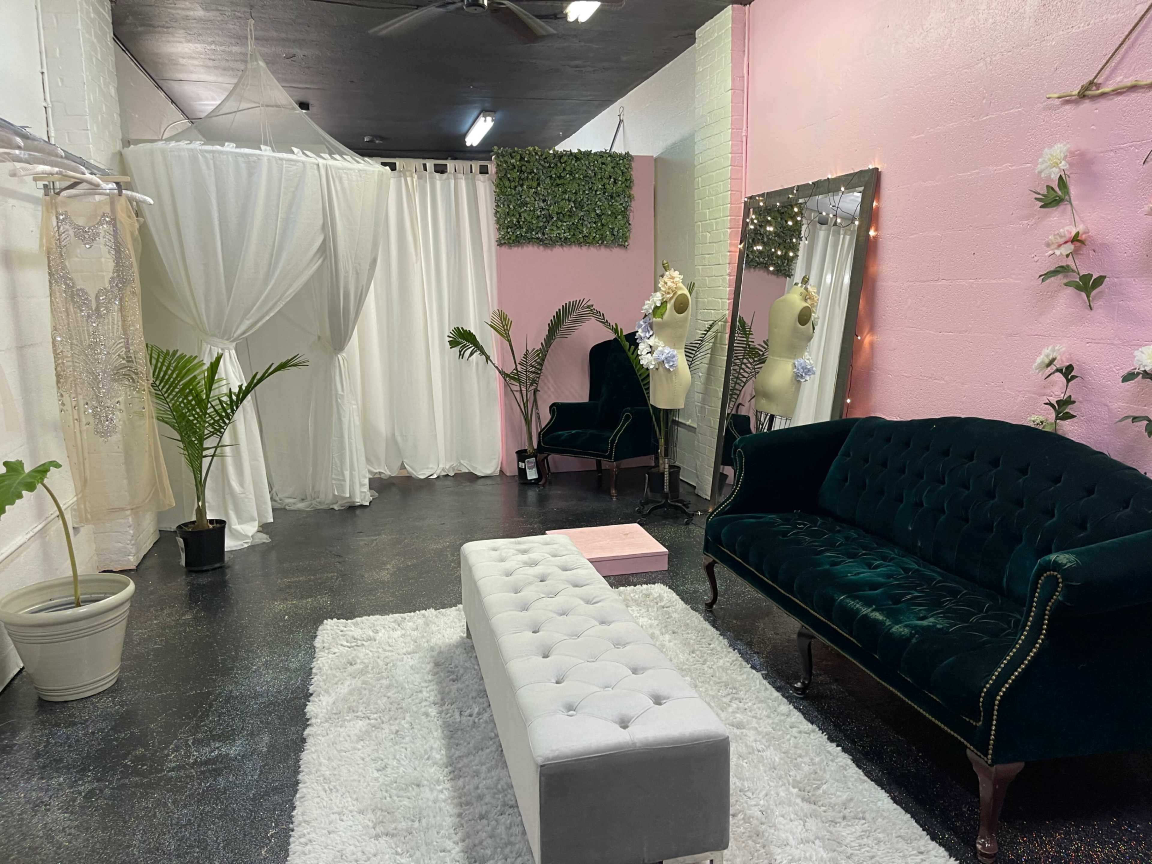 A room featuring a green velvet sofa, a white bench, and draped fabric partitions, with decorative plants and a large mirror.