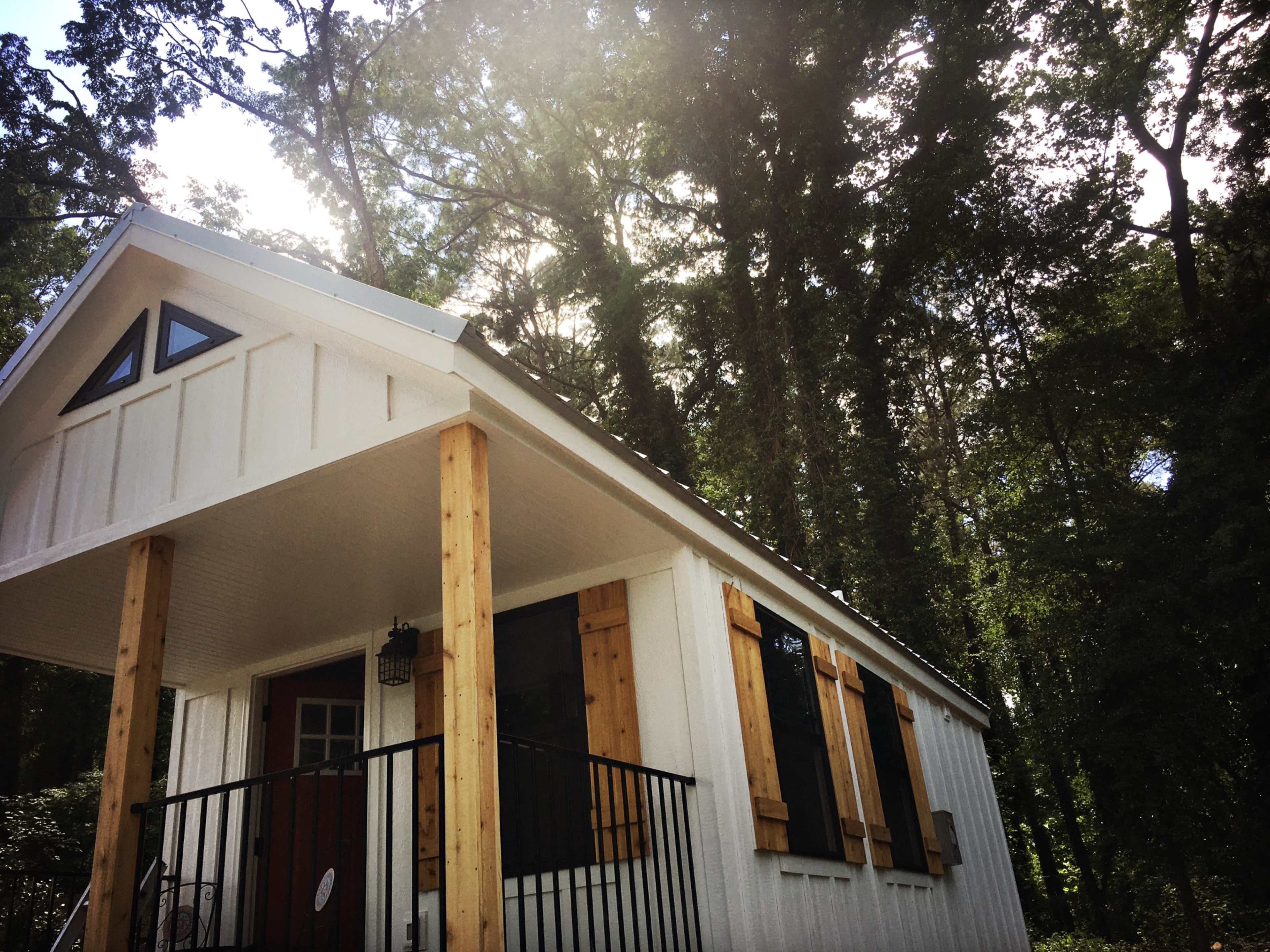 A small white house with wooden shutters and a porch stands among tall trees.