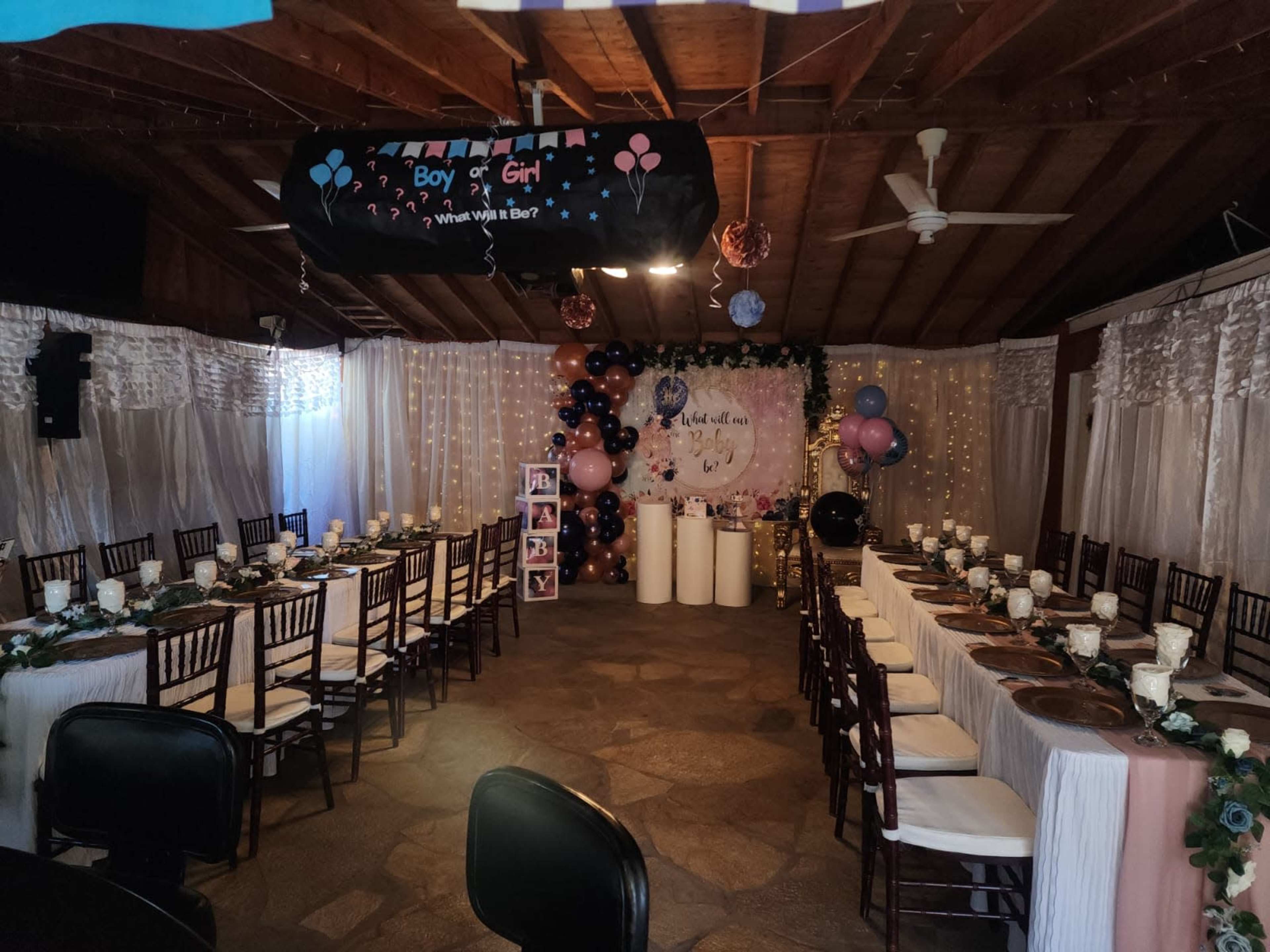 A decorated indoor venue for a gender reveal party, featuring long tables set with white tablecloths, centerpieces, and balloons.