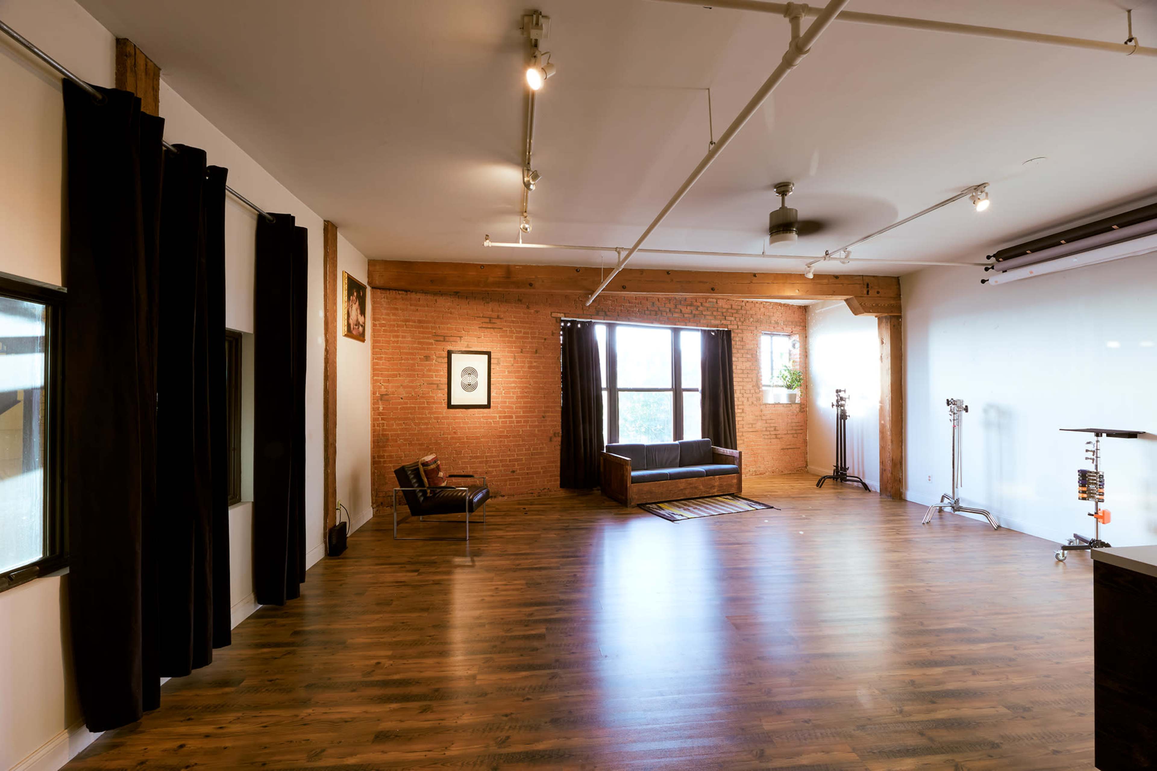 The image shows a spacious studio with wooden floors, exposed brick walls, large windows, and minimalist furnishings.