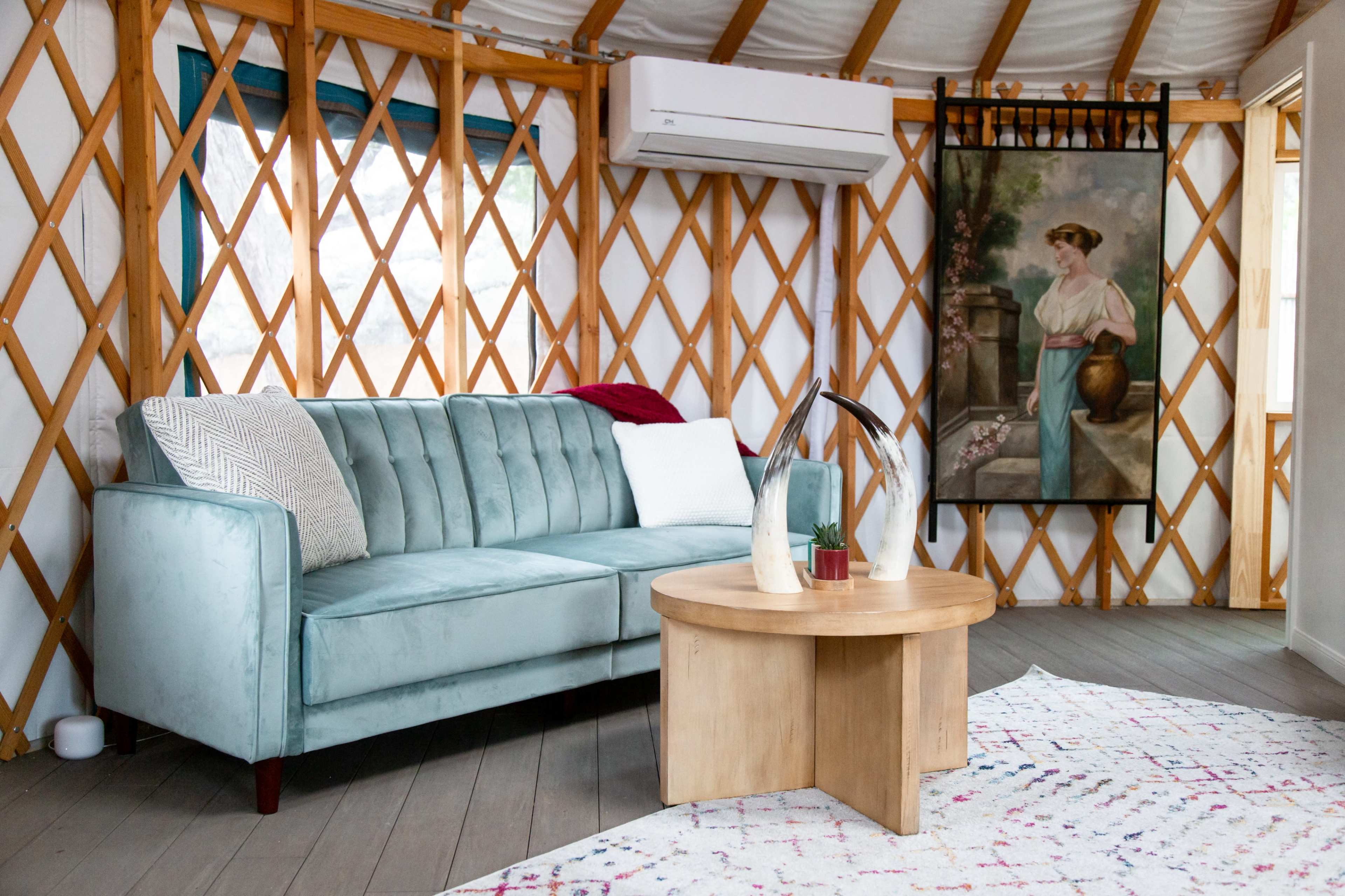 A cozy yurt interior featuring a light blue couch, a wooden round coffee table, and a large framed painting of a woman next to a window.