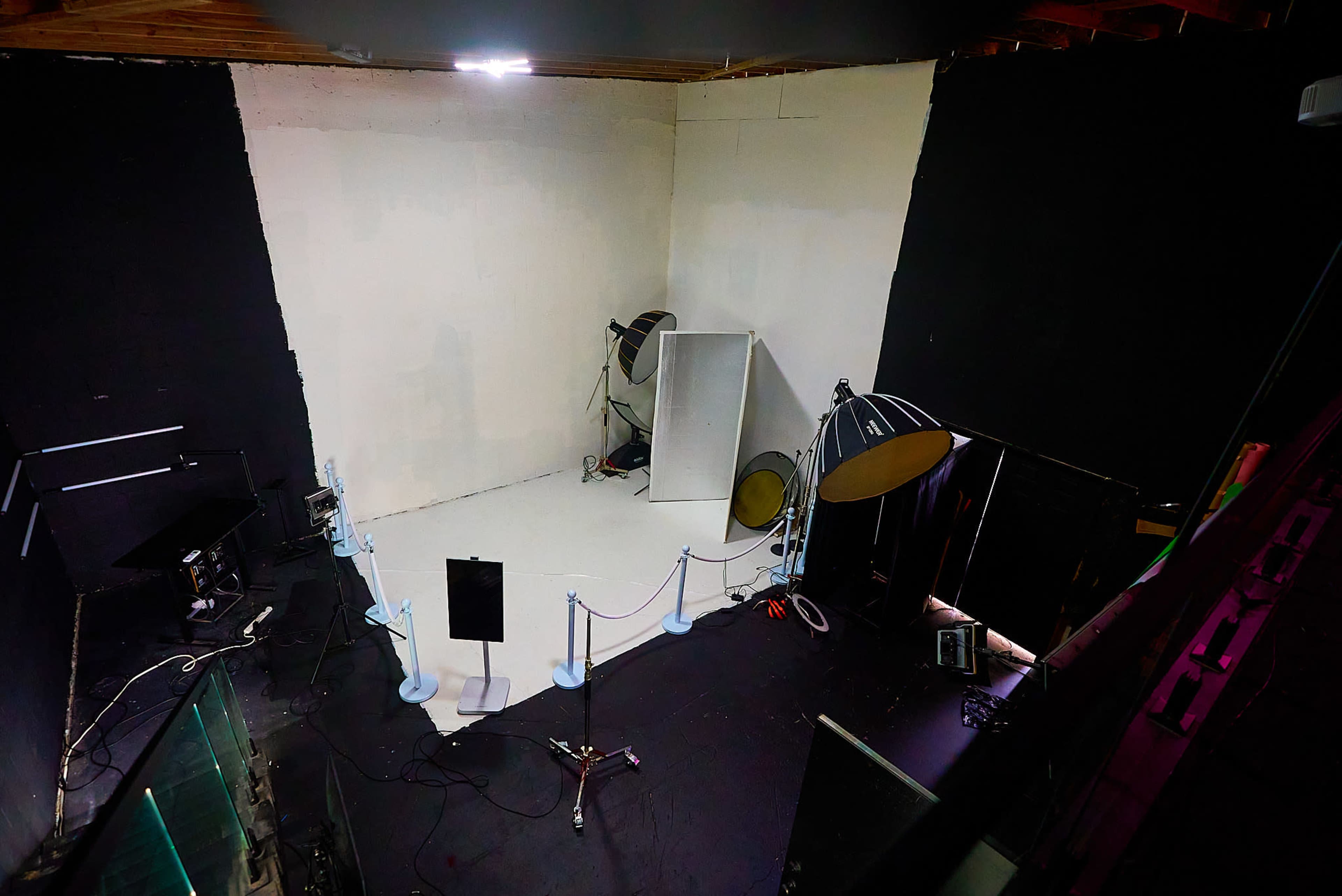 The image shows a photo studio featuring a blank white wall, various lighting equipment, and a small sectioned area marked off with a rope.