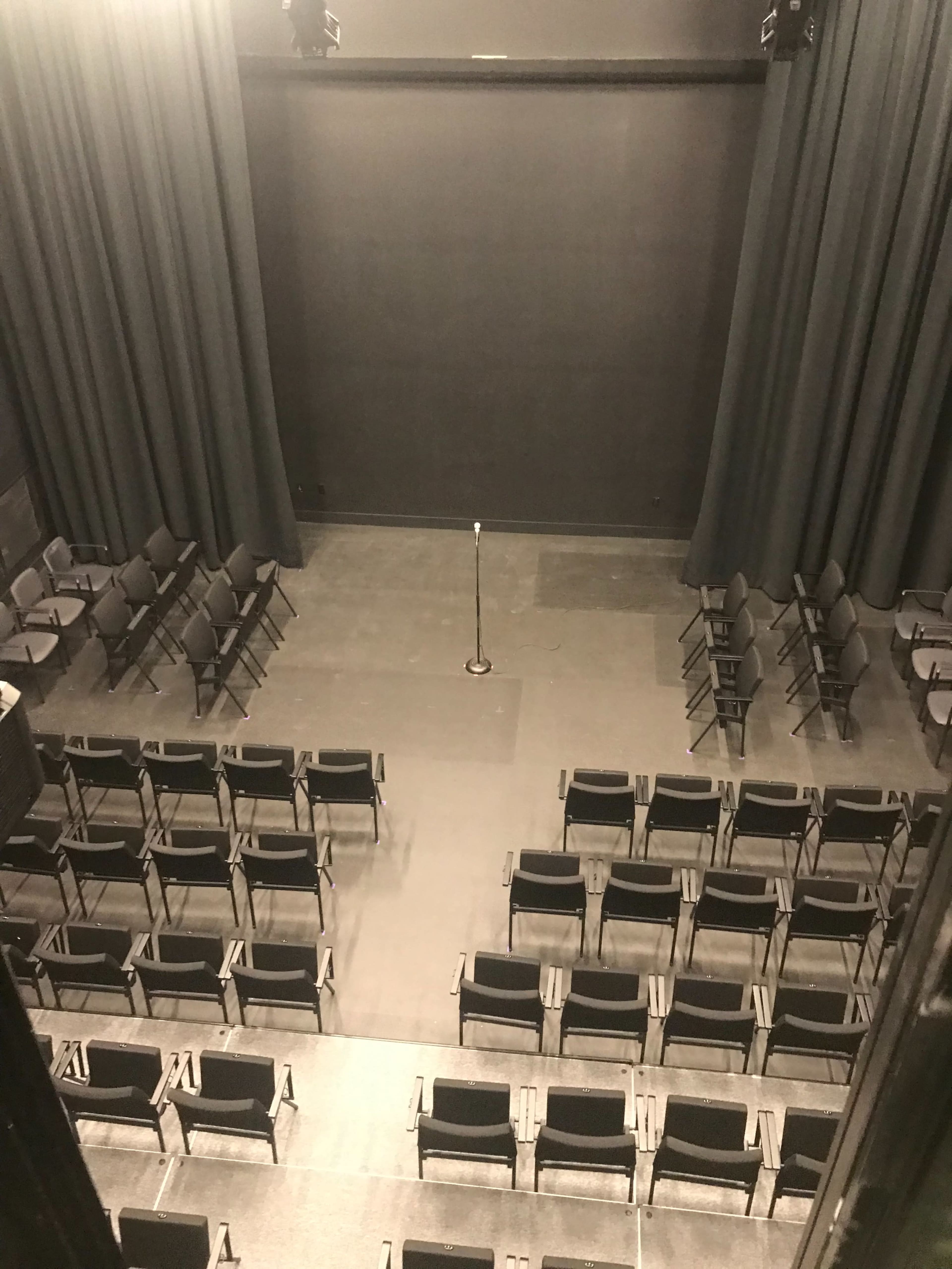 A theater stage with a microphone stand in the center and rows of empty chairs arranged in front of it.