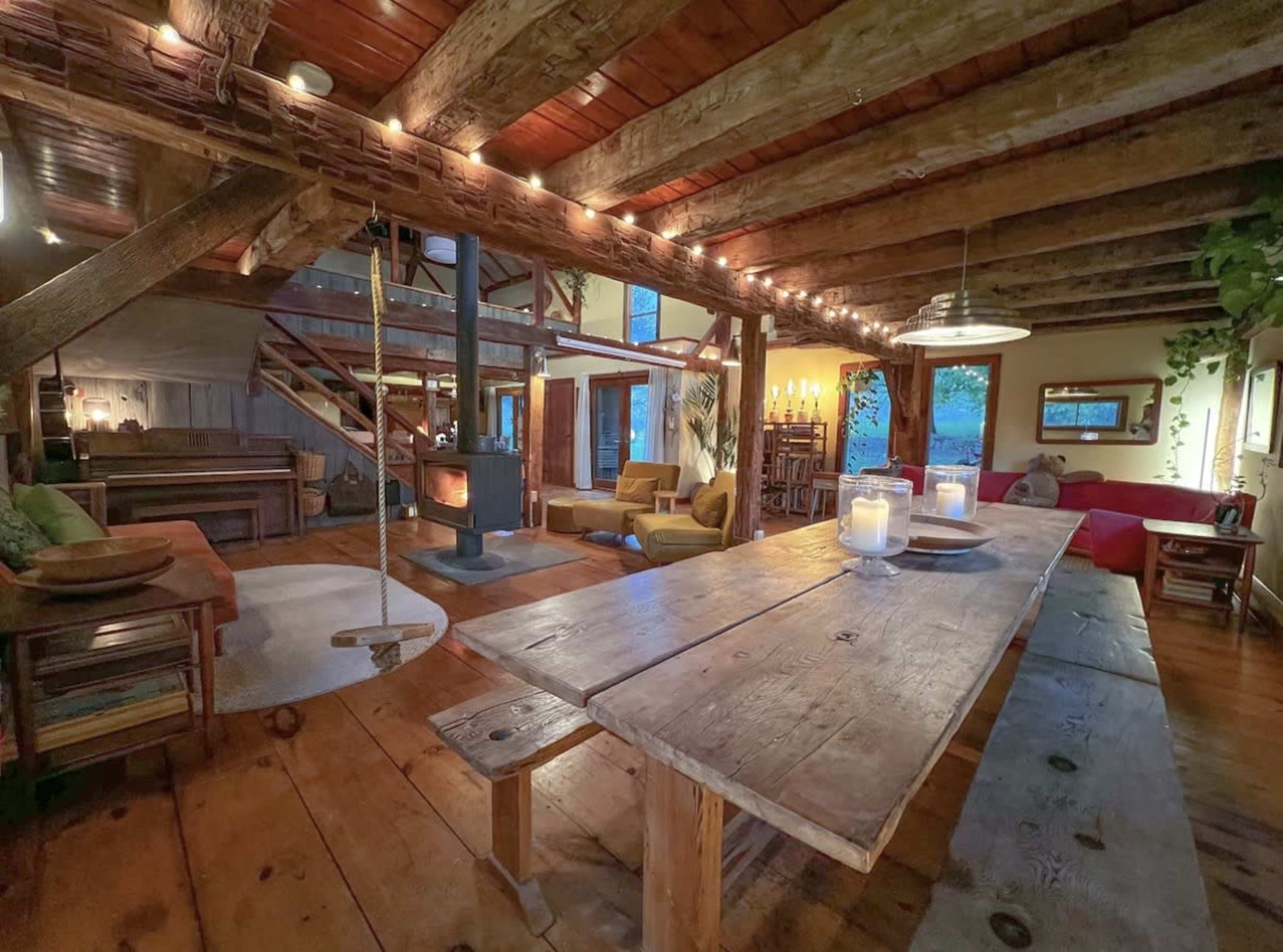 The image shows an inviting rustic interior of a house featuring wooden beams, a large dining table, cozy seating areas, and warm lighting.