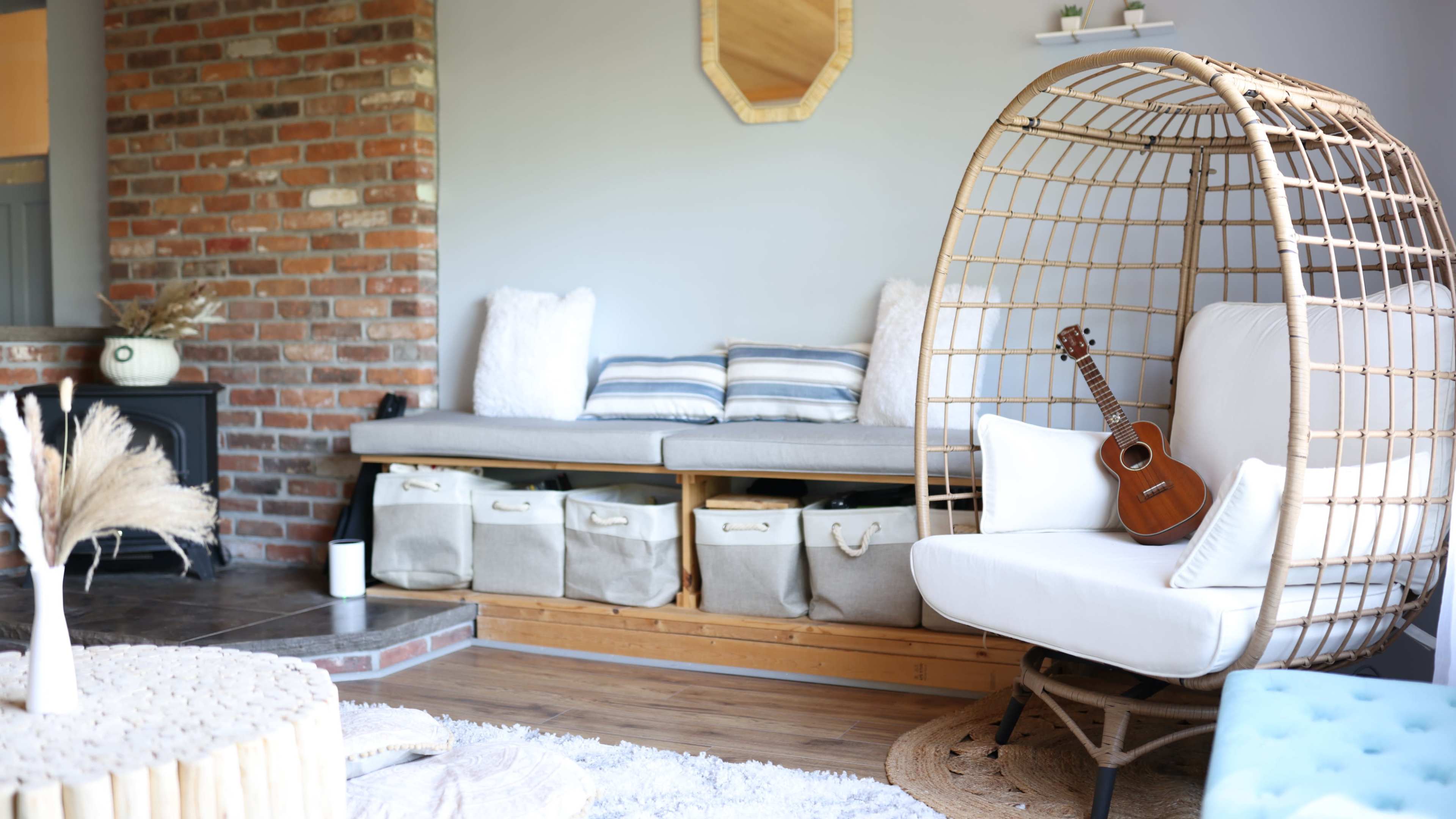 A cozy living room with a rattan chair, a small ukulele resting on it, and a sofa with storage baskets underneath.