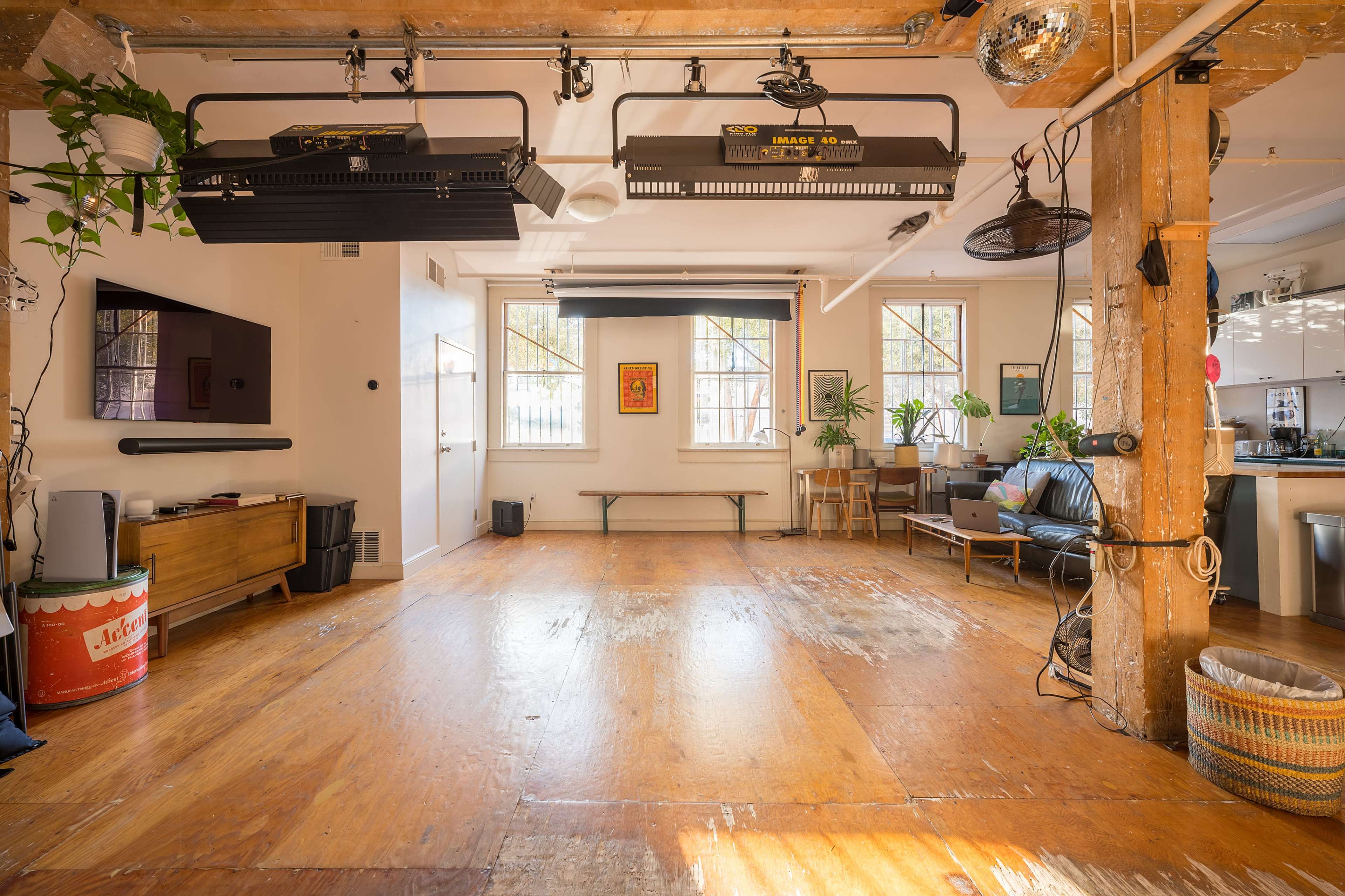An open room with wooden flooring, light fixtures mounted on the ceiling, potted plants, a black sofa, and a wooden bench along with a kitchen area visible in the background.