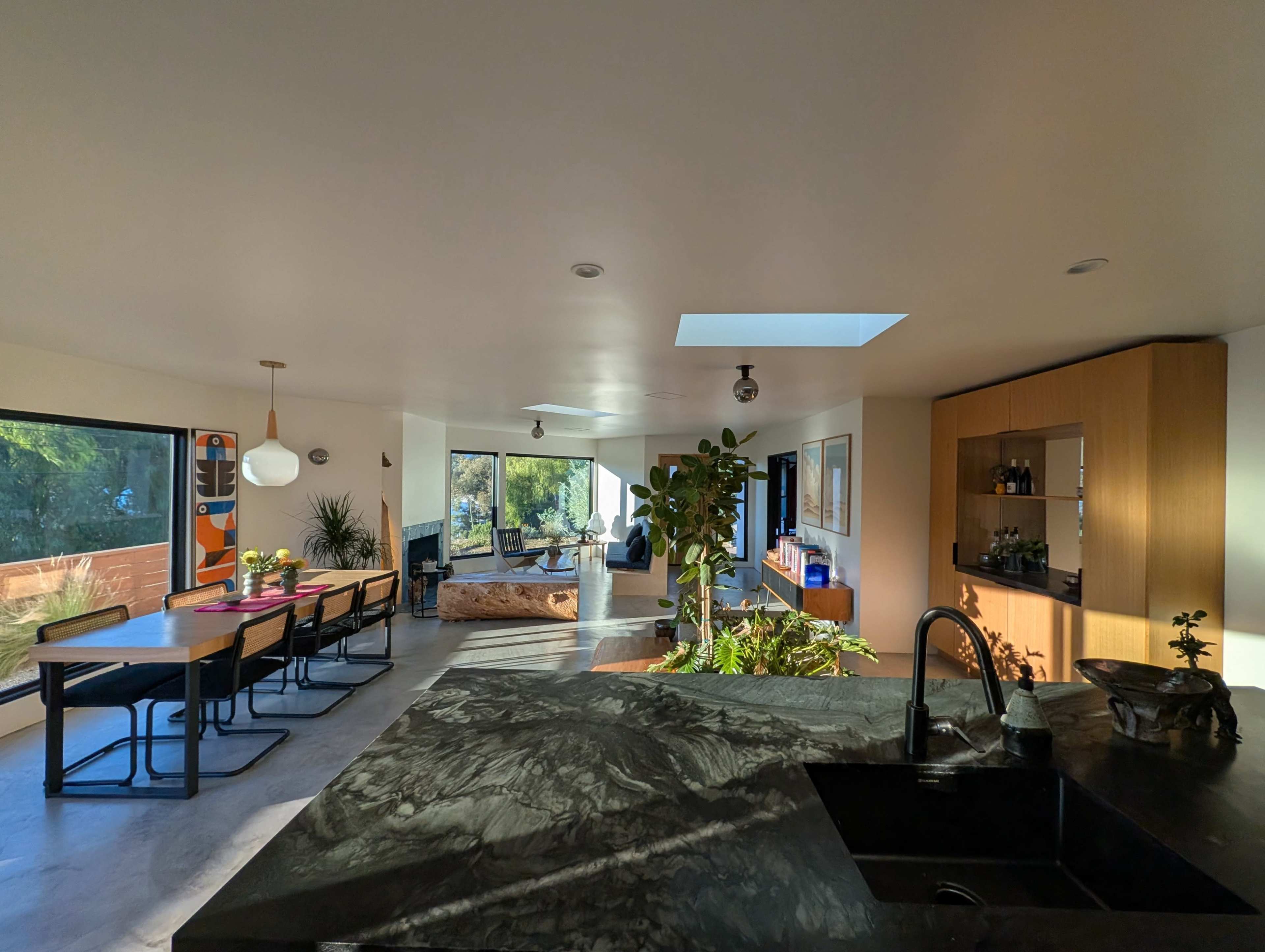 The image shows a modern open-concept living space featuring a kitchen, dining area, and lounge with large windows and natural light.