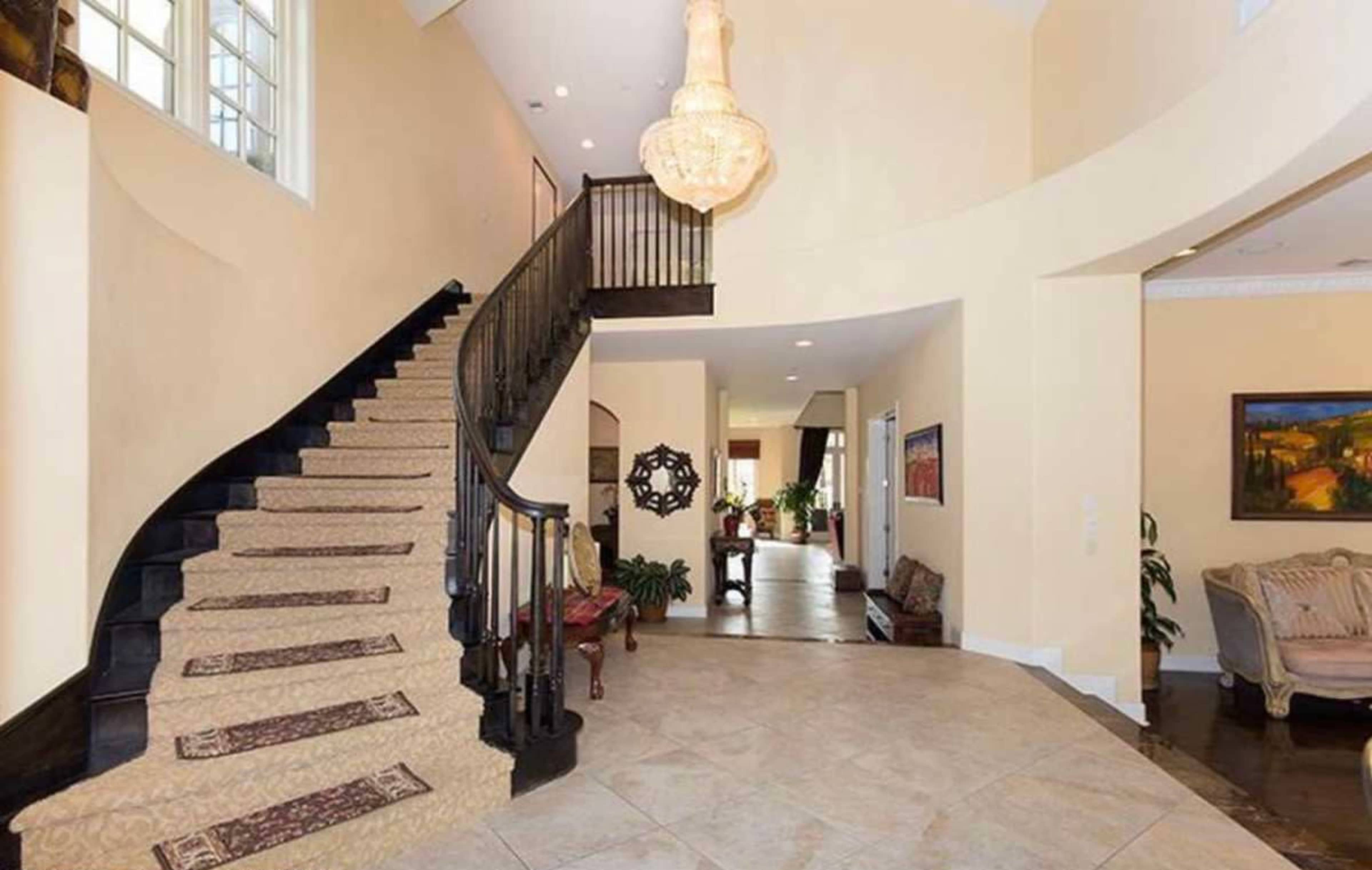 The image shows a spacious foyer with a curved staircase, a chandelier overhead, and various decorative elements, including seating and plants.