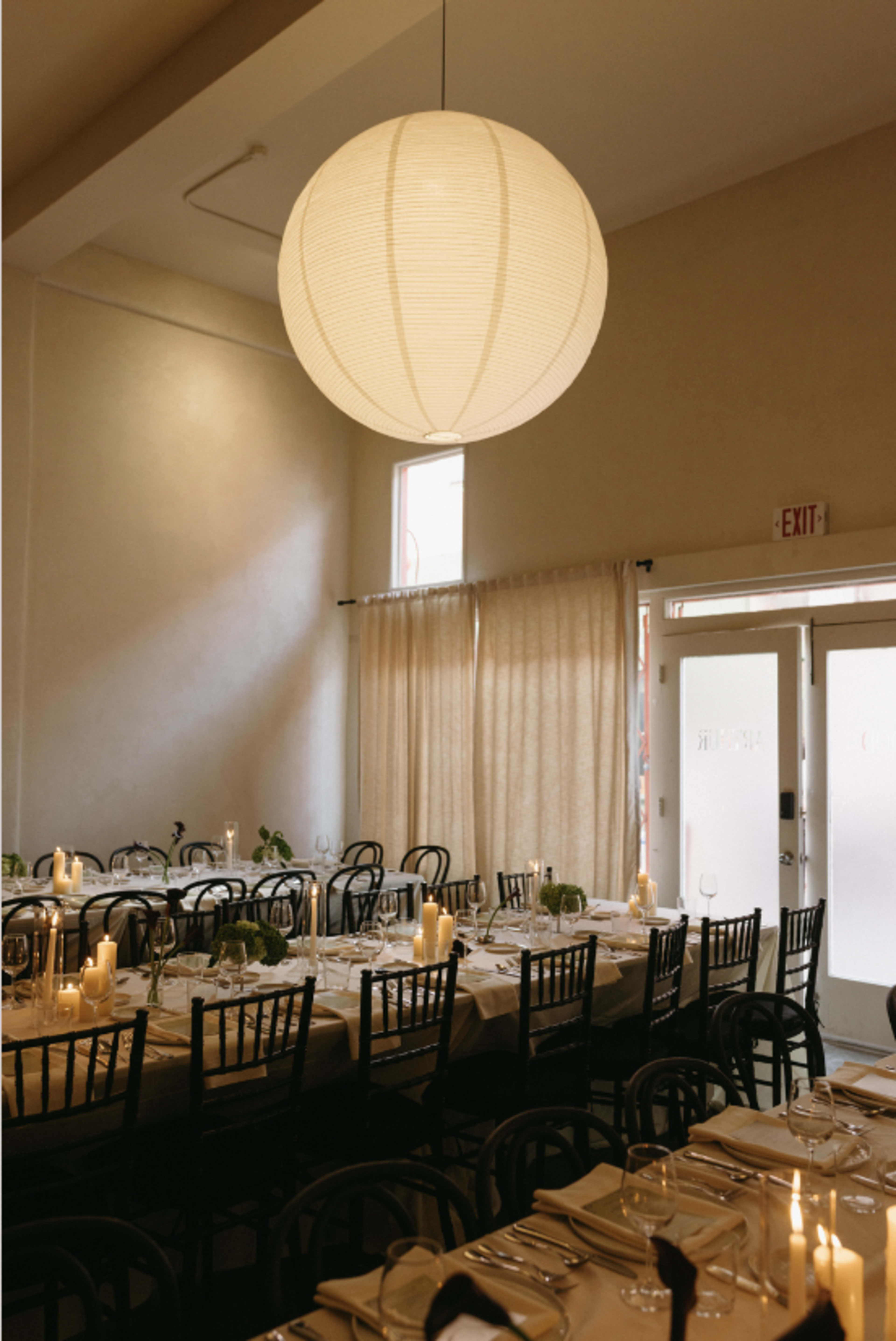 A large, round paper lantern hangs above a long dining table set with candles and plates in a softly lit restaurant interior.