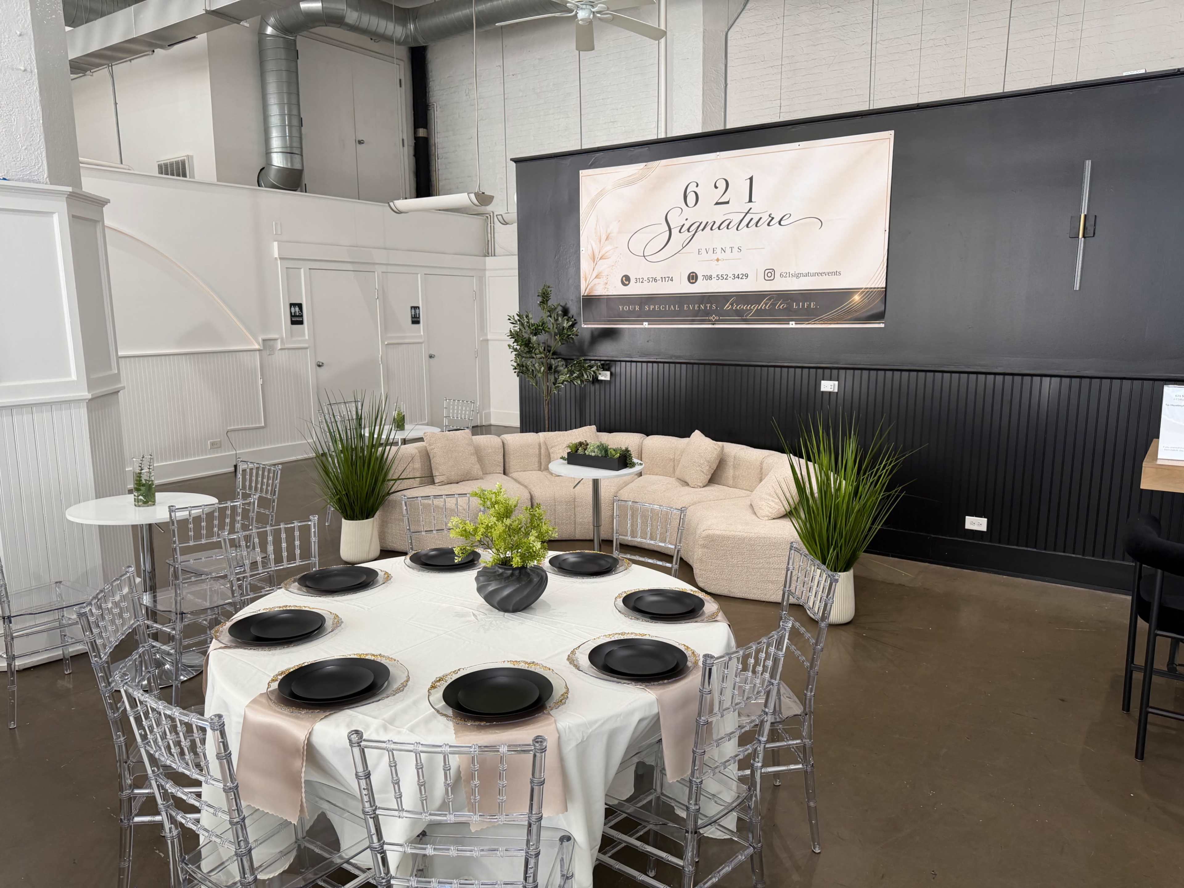 The image shows a modern event space with a round table set for dining, featuring black plates, a light-colored tablecloth, and a large, L-shaped sectional sofa along a black wall adorned with a promotional banner.
