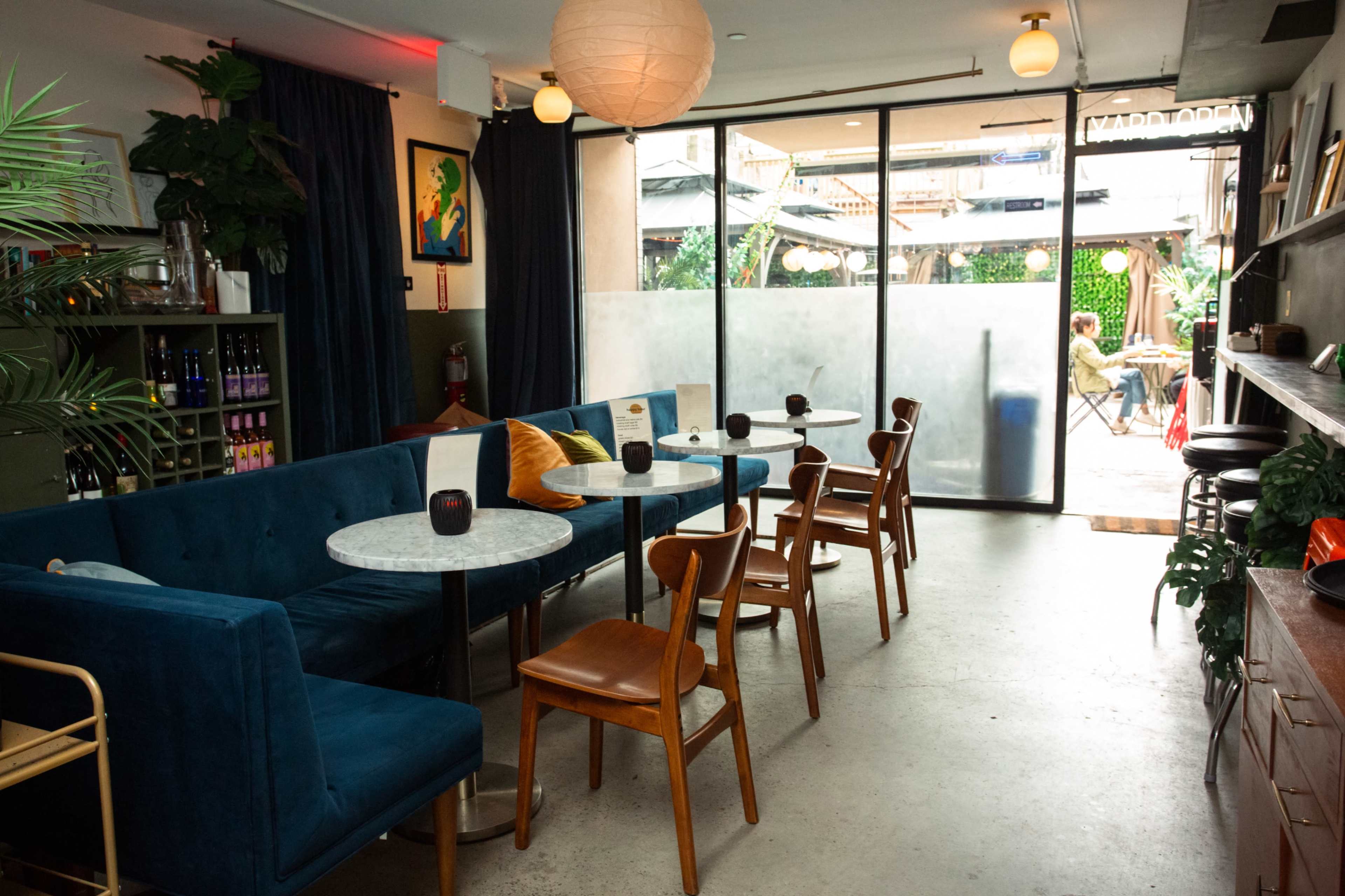 A cozy cafe interior with blue velvet seating, wooden chairs, and a view of an outdoor seating area through large glass doors.