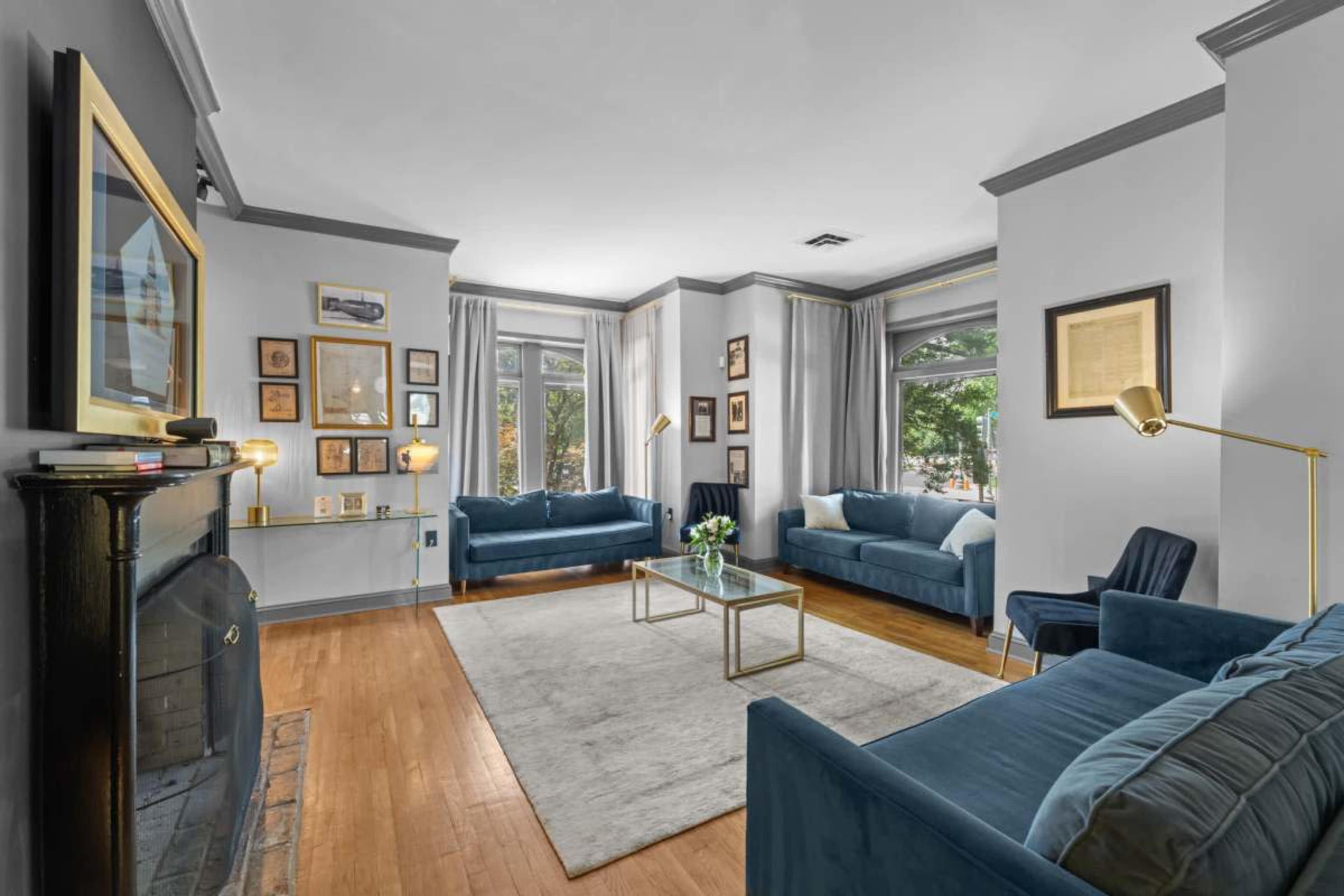 A spacious living room features two blue sofas, a coffee table, and various framed artworks on the walls, illuminated by natural light from the windows.