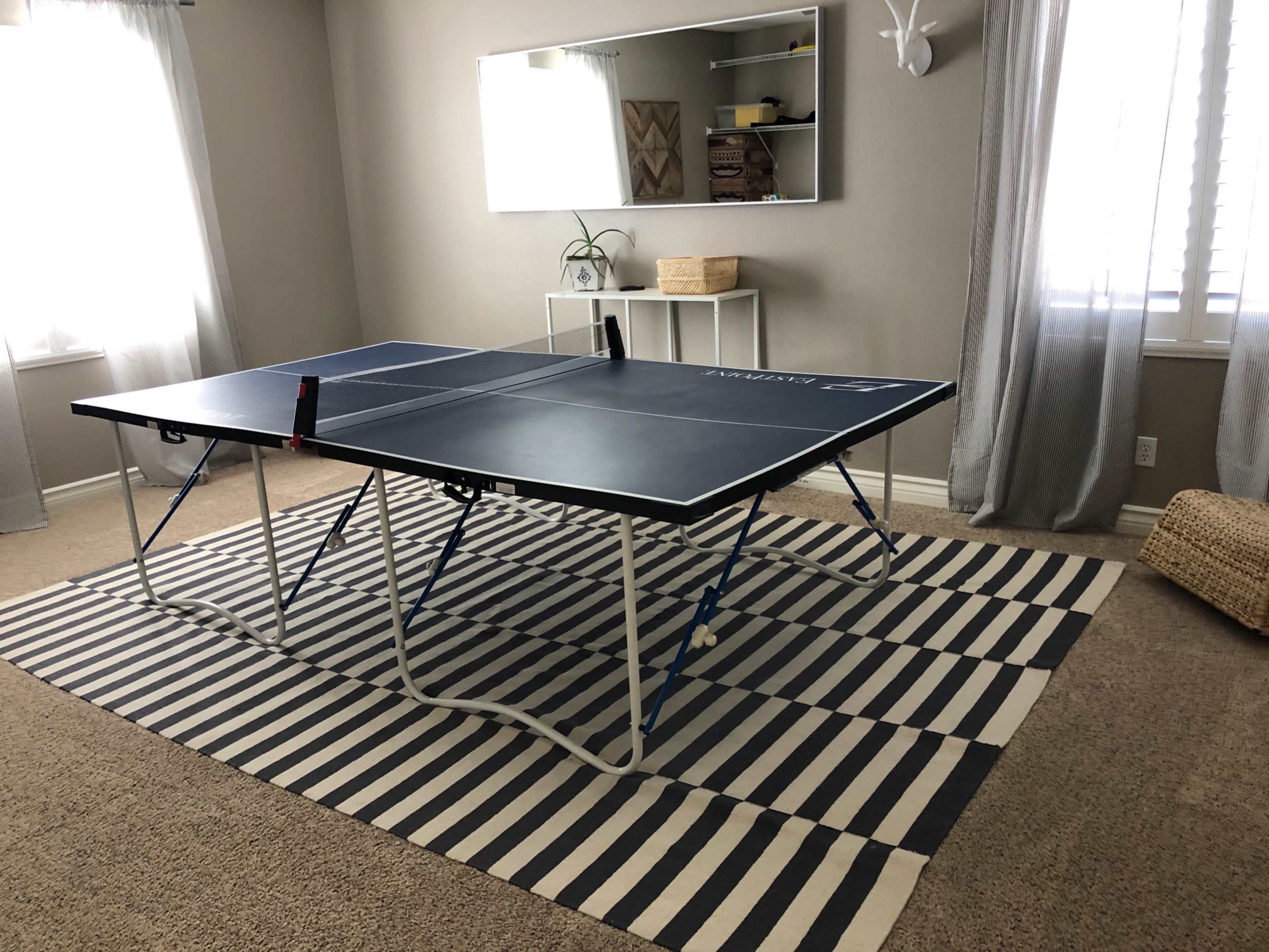 A ping pong table is set up in a living room with a striped rug and a large mirror on the wall.