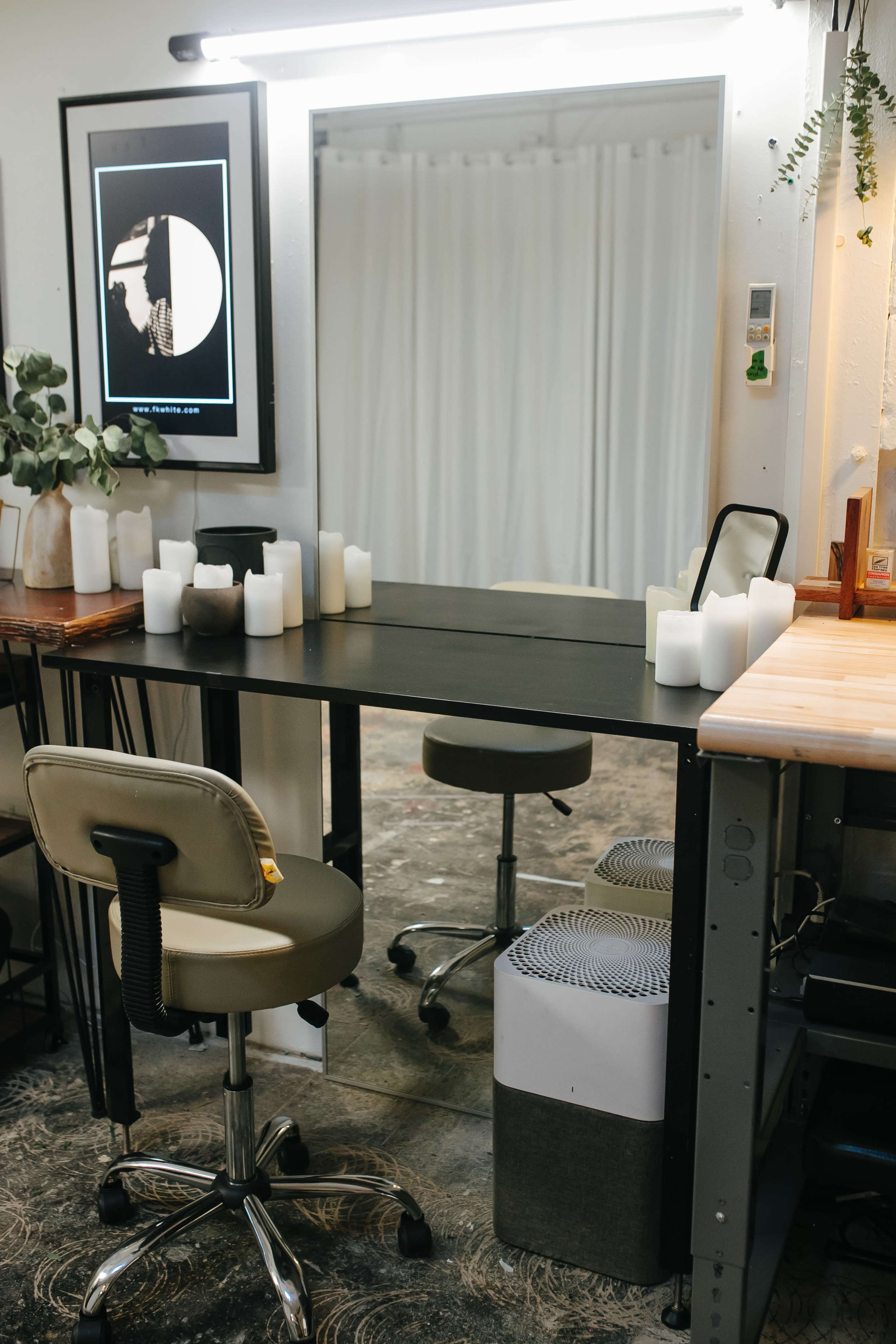 A workspace features a black table with a large mirror above it, surrounded by several white candles and two rolling office chairs.