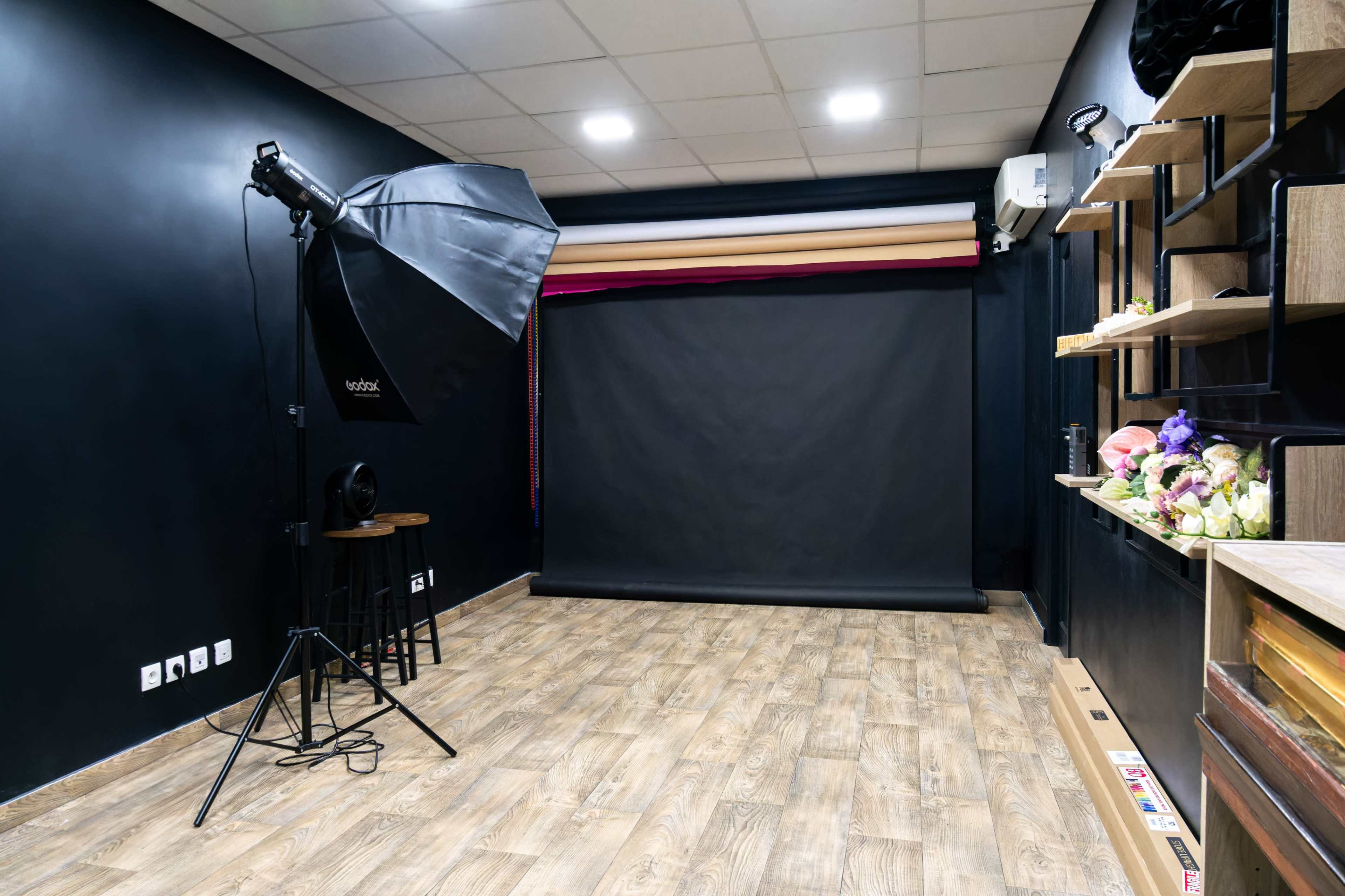 The image shows a photography studio with a black backdrop, a large softbox light on a stand, and wooden flooring.