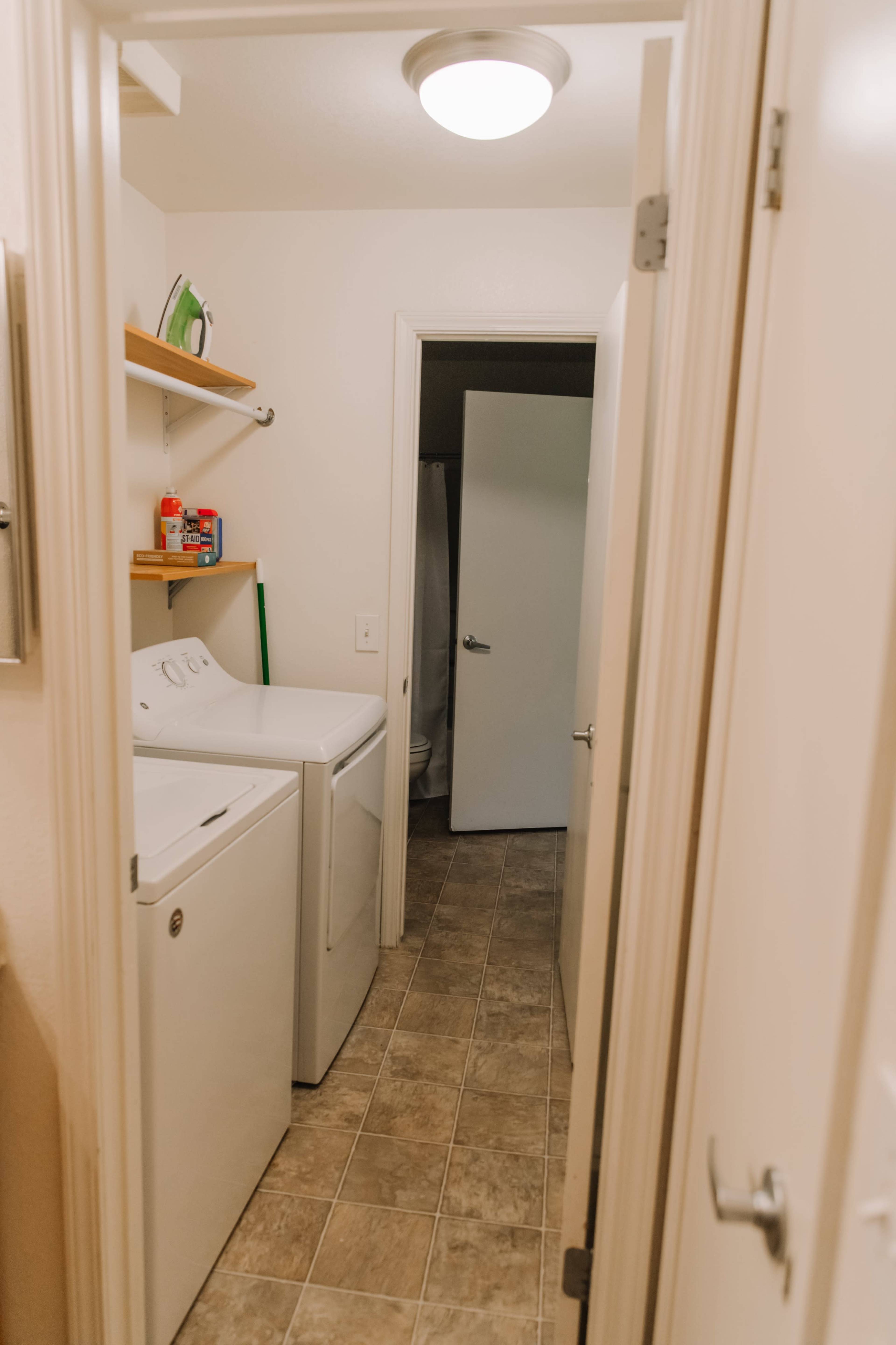 A laundry room features a washer and dryer against one wall, with shelves above them and a closed door at the end of the hallway.