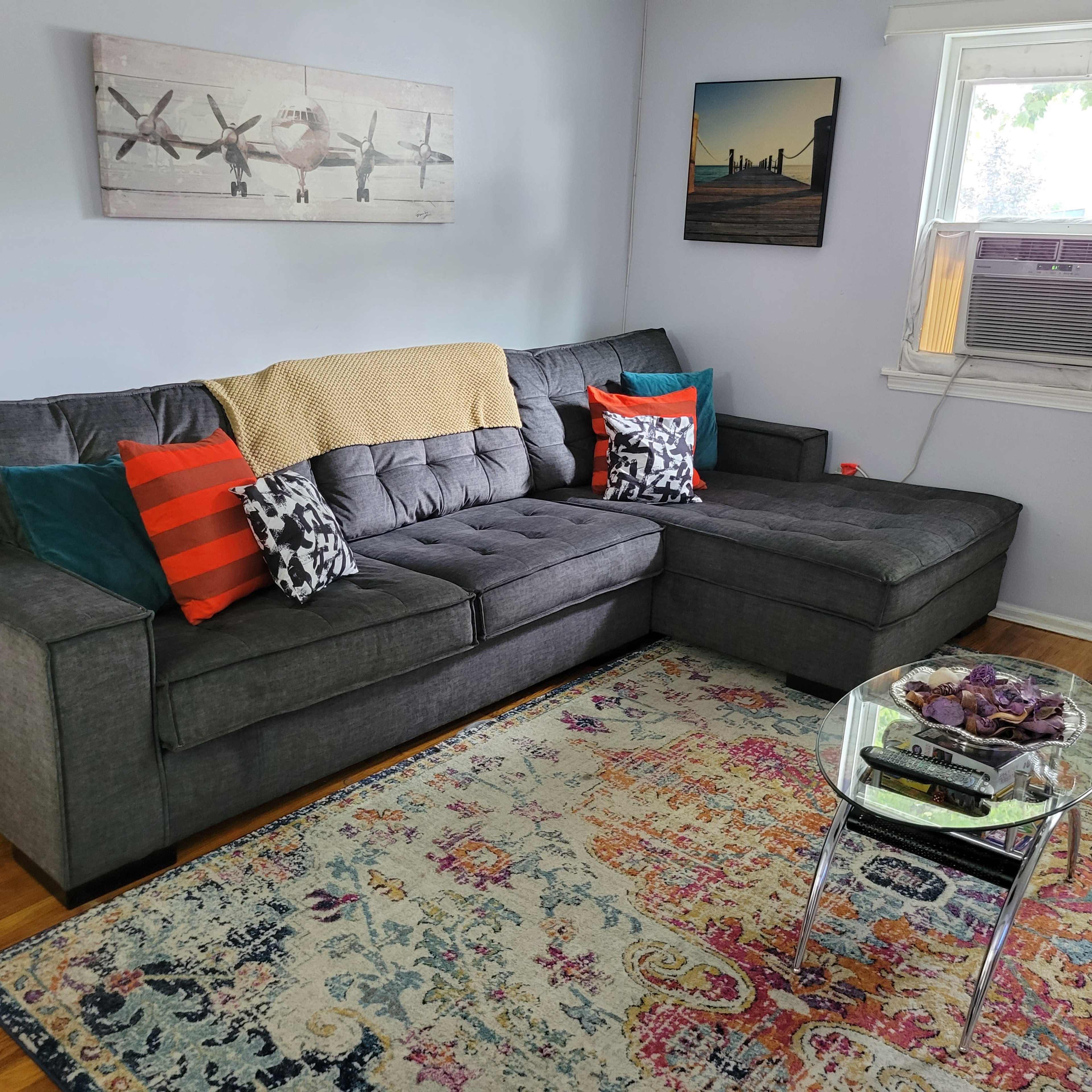 A gray sectional sofa with colorful cushions sits on a patterned rug in a bright room featuring a wall-mounted air conditioner and a window.