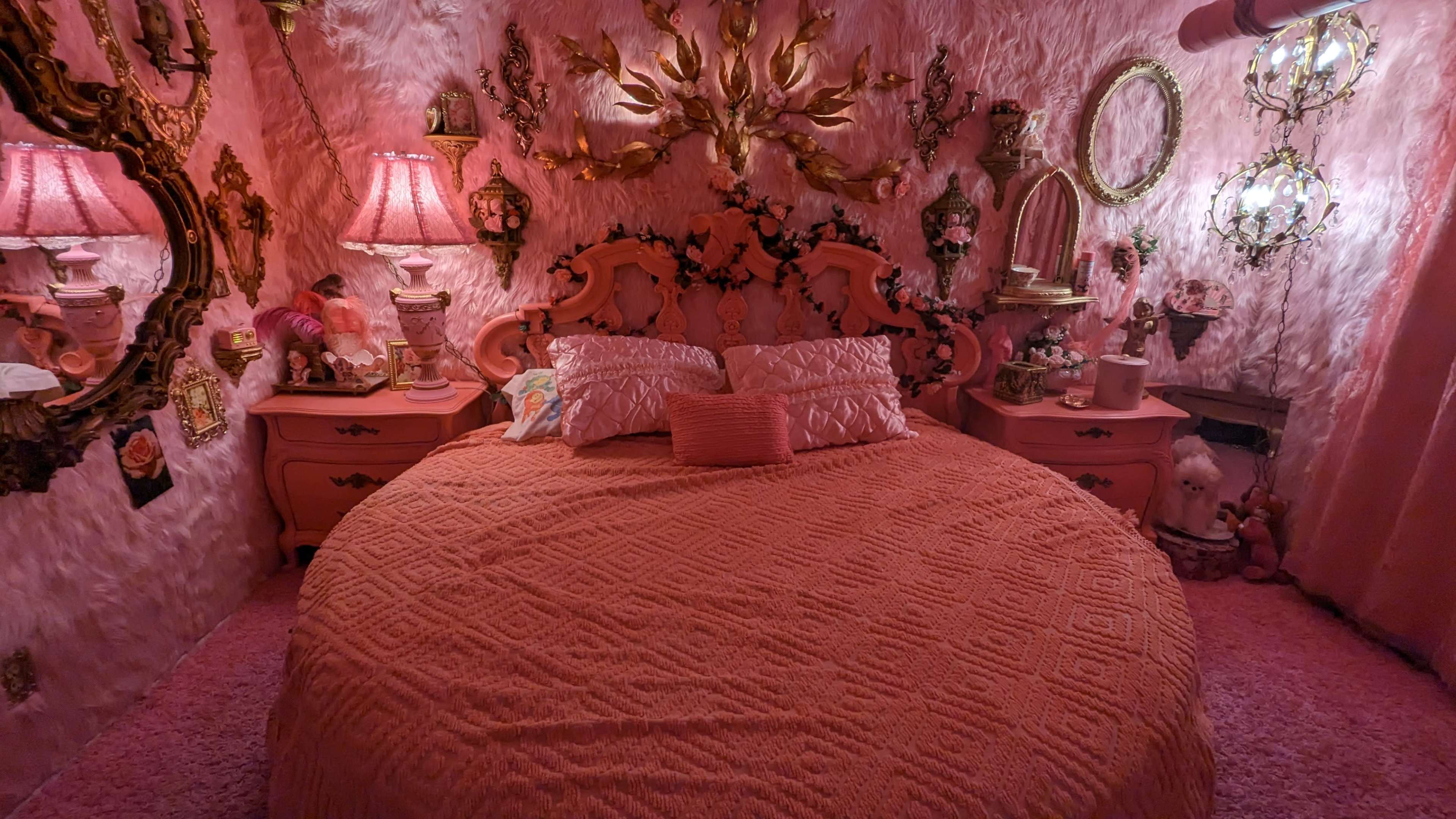 The bedroom features a round bed with a textured pink coverlet, surrounded by walls adorned with pink faux fur and decorative mirrors, lamps, and wall art.