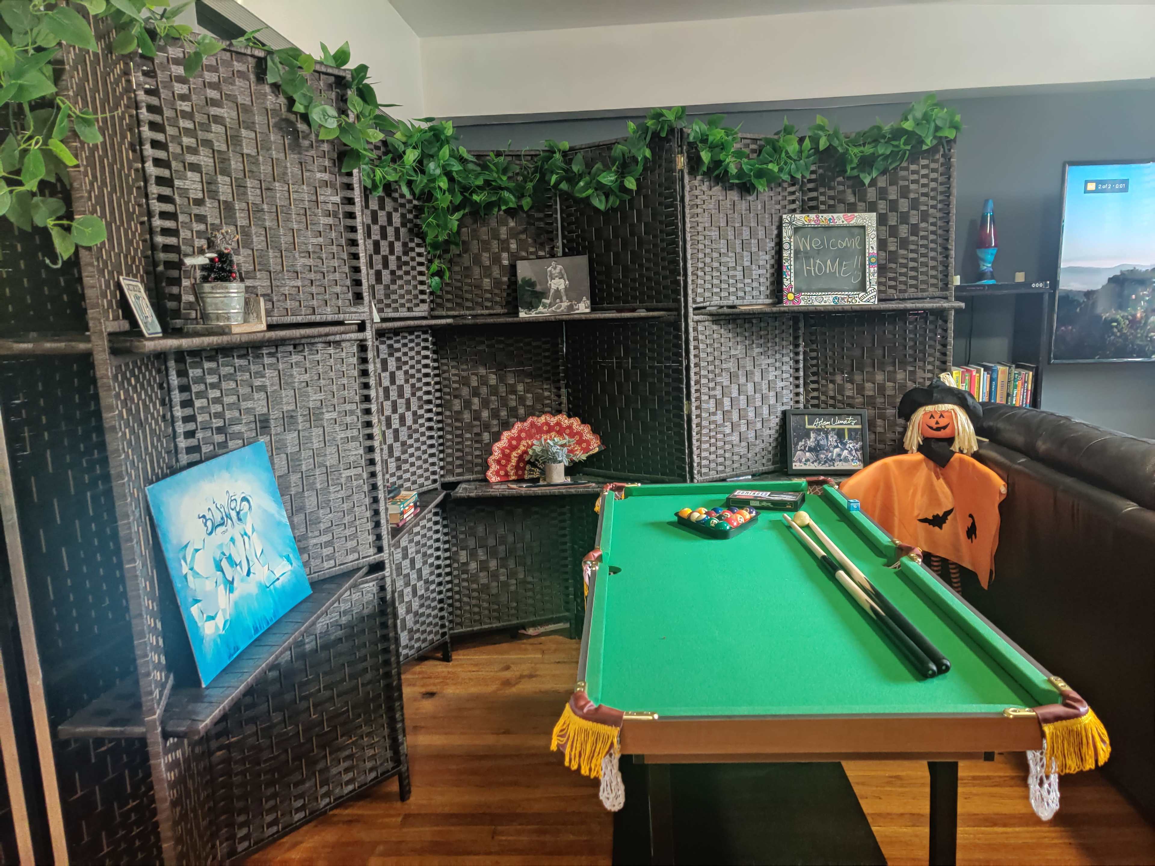 A room with a green pool table in the center, surrounded by black woven shelves and decorated with plants, a scarecrow, and various framed pictures.