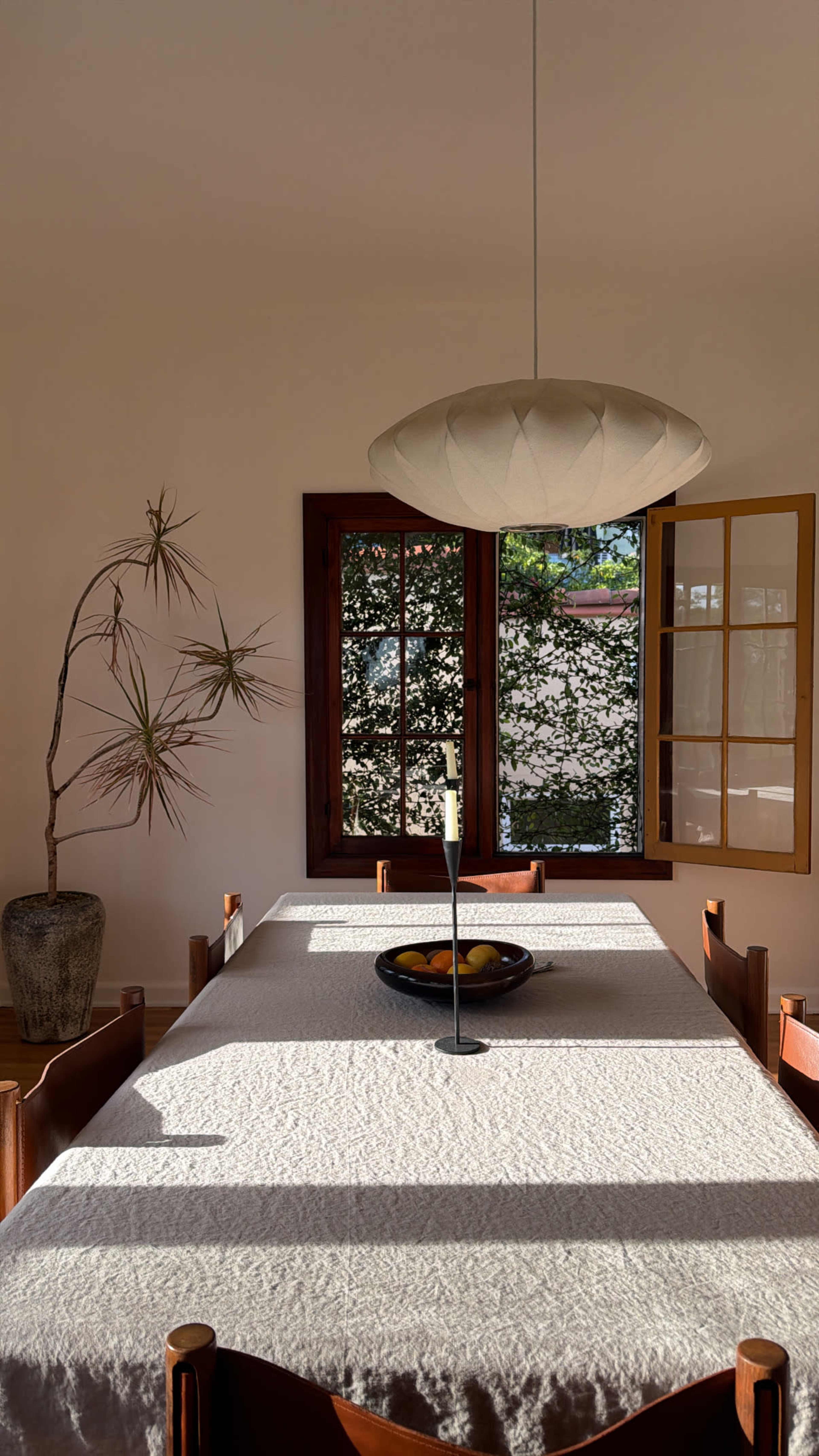 A dining table with a textured cloth is set in a well-lit room, featuring a central candle holder, a bowl of fruit, and a potted plant by a window.