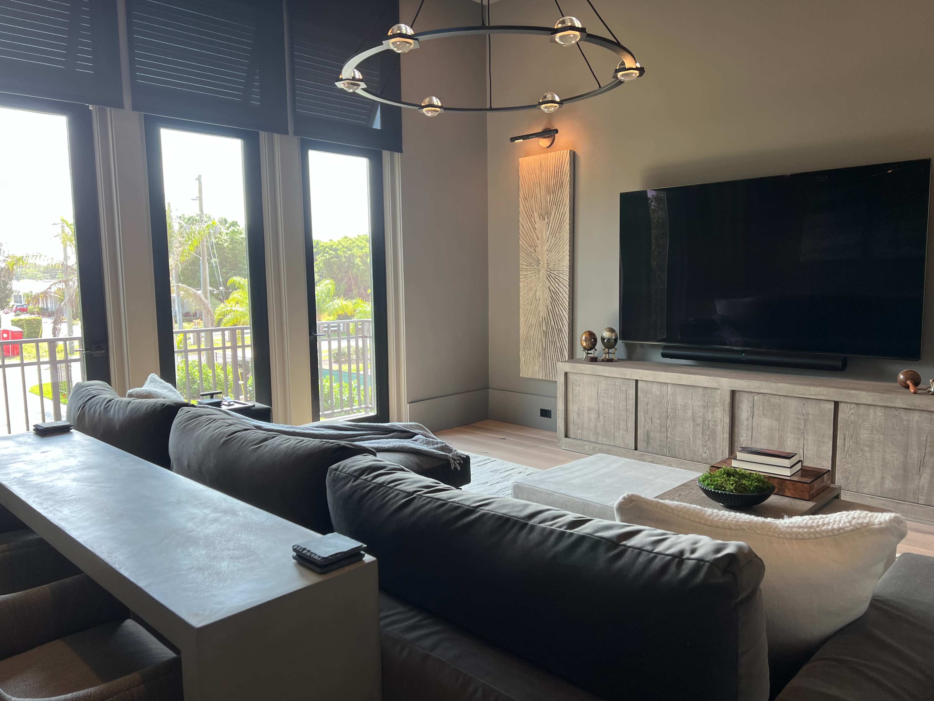 A modern living room with a large television mounted on the wall, a sectional sofa, and windows overlooking a view outside.