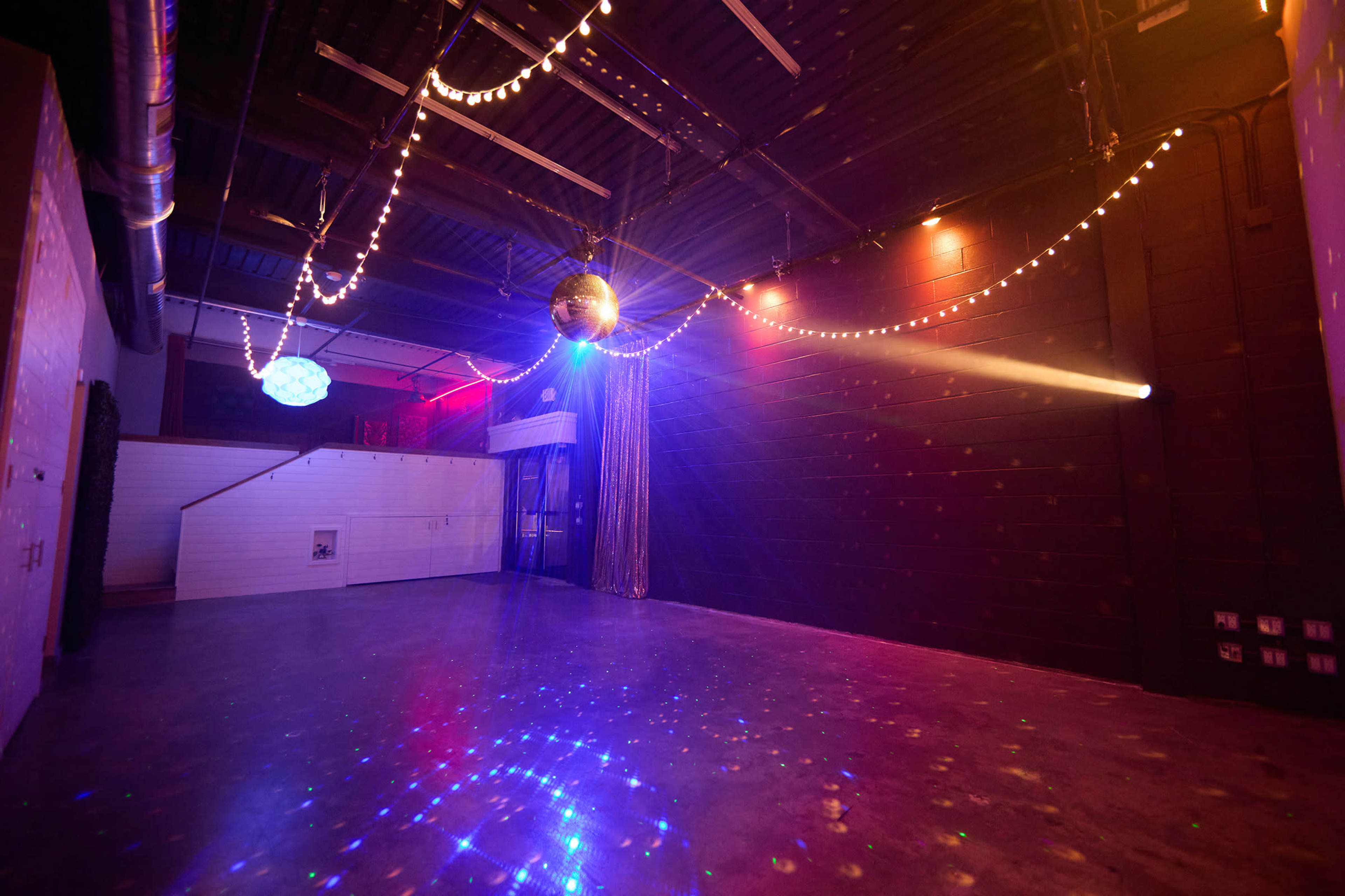 The image shows an empty event space with a polished concrete floor, decorated with string lights and a disco ball reflecting colorful lights against the walls.