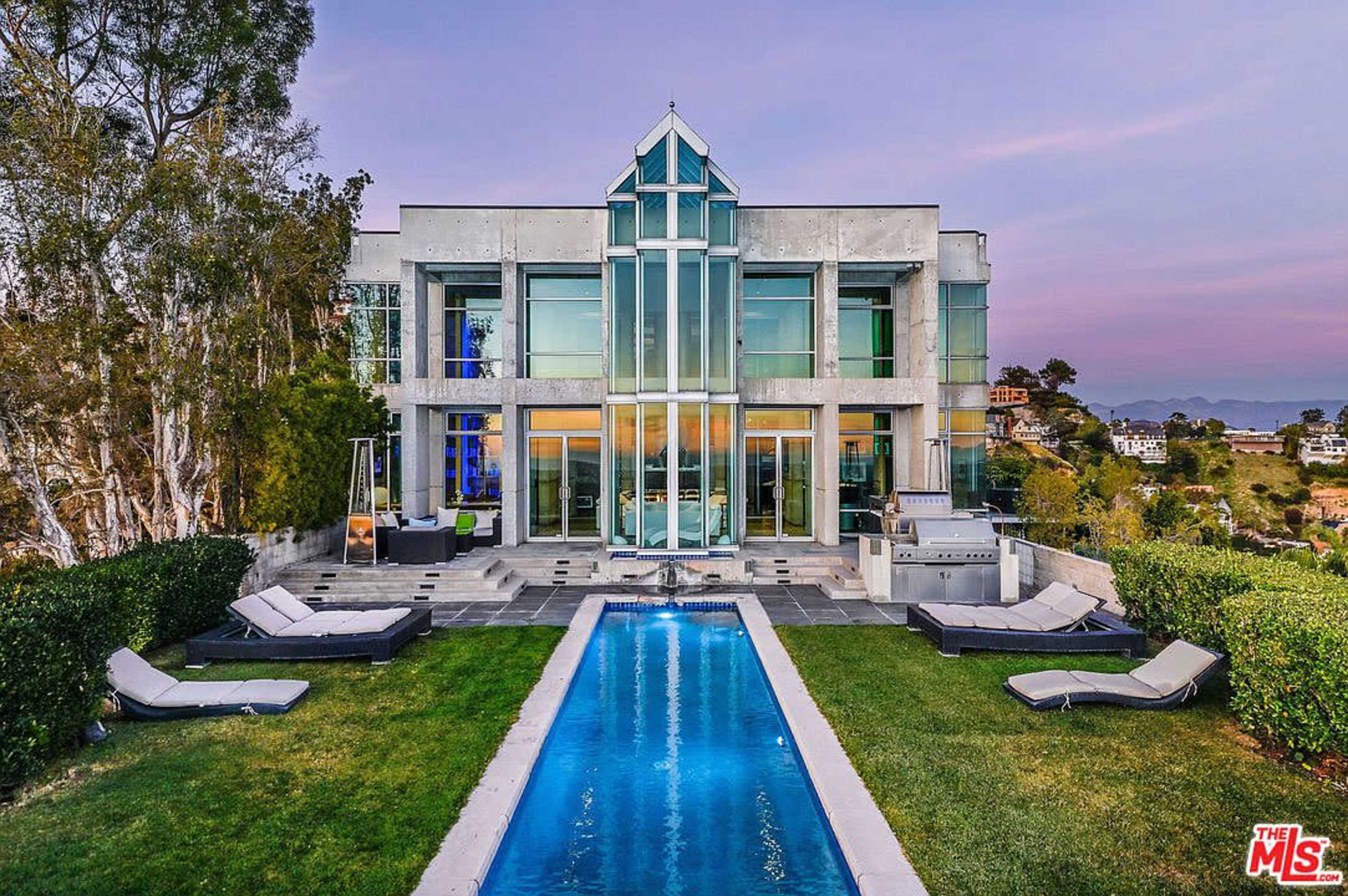 The image shows a modern two-story house with large glass windows, a swimming pool, and lounge chairs in a landscaped backyard.