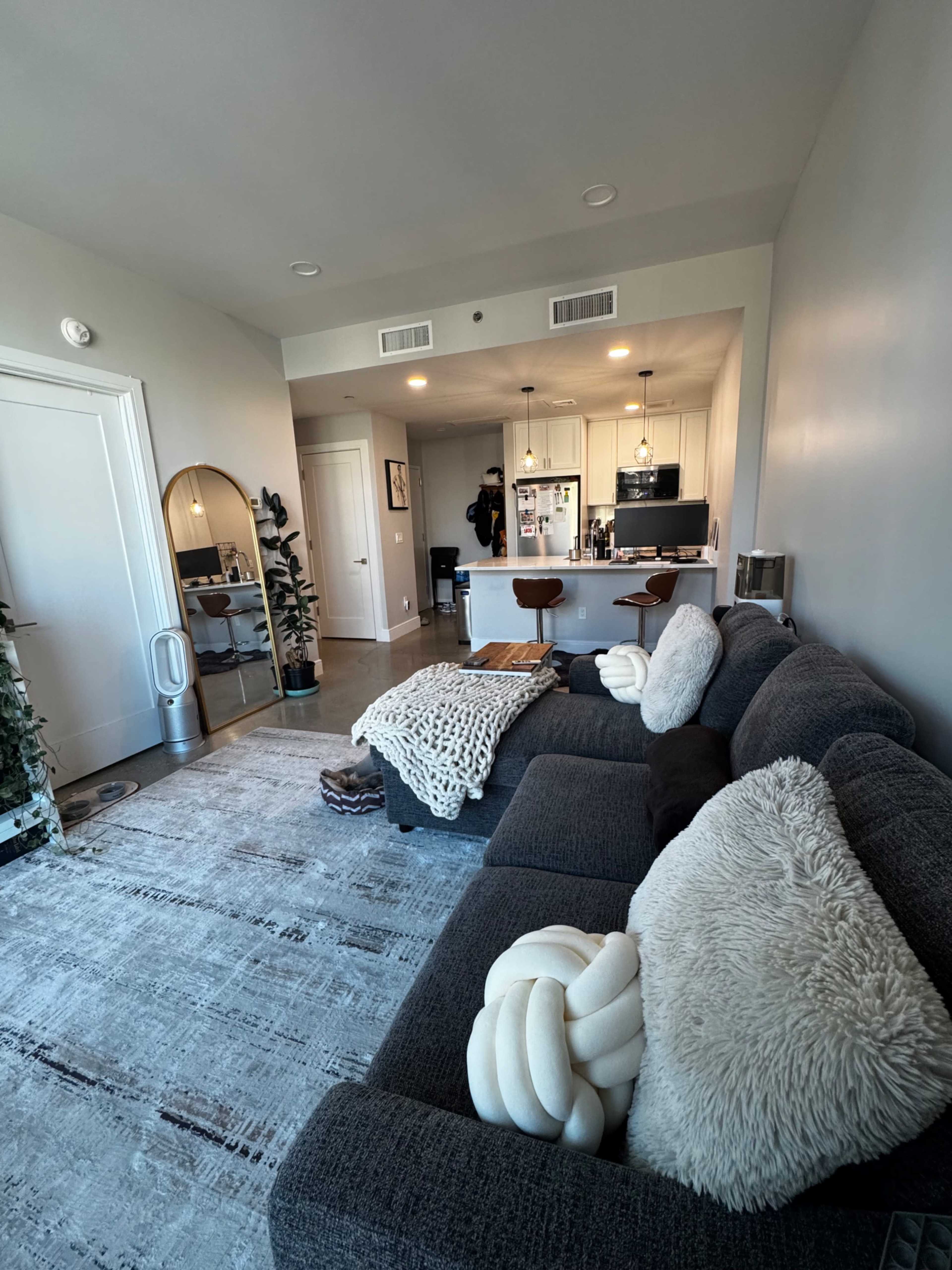 The image shows a modern living room featuring a gray sectional sofa, a coffee table with decorative items, and a view into a kitchen area with bar stools and appliances.
