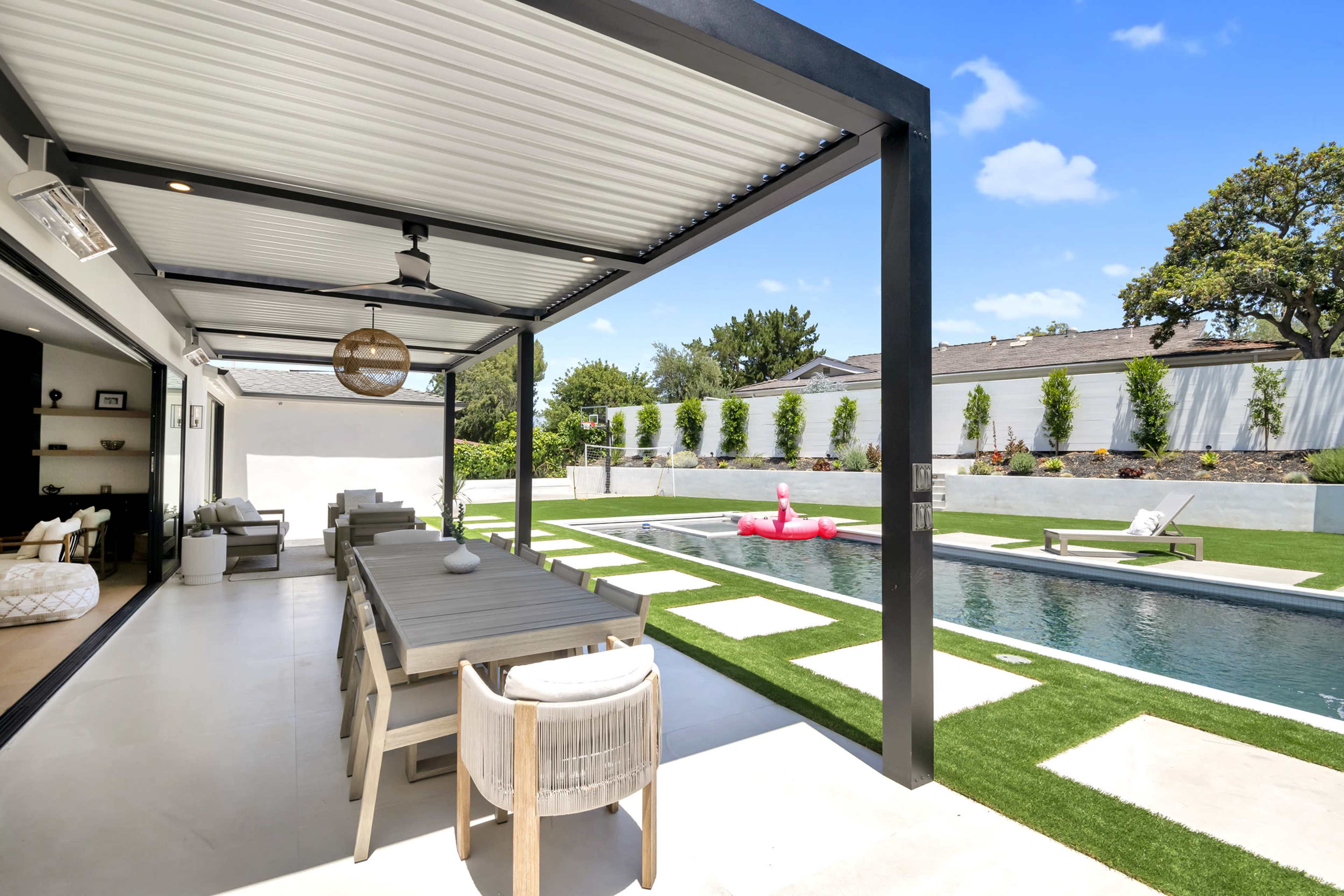 A covered patio area features a long dining table overlooking a landscaped backyard with a swimming pool and lounge chairs.