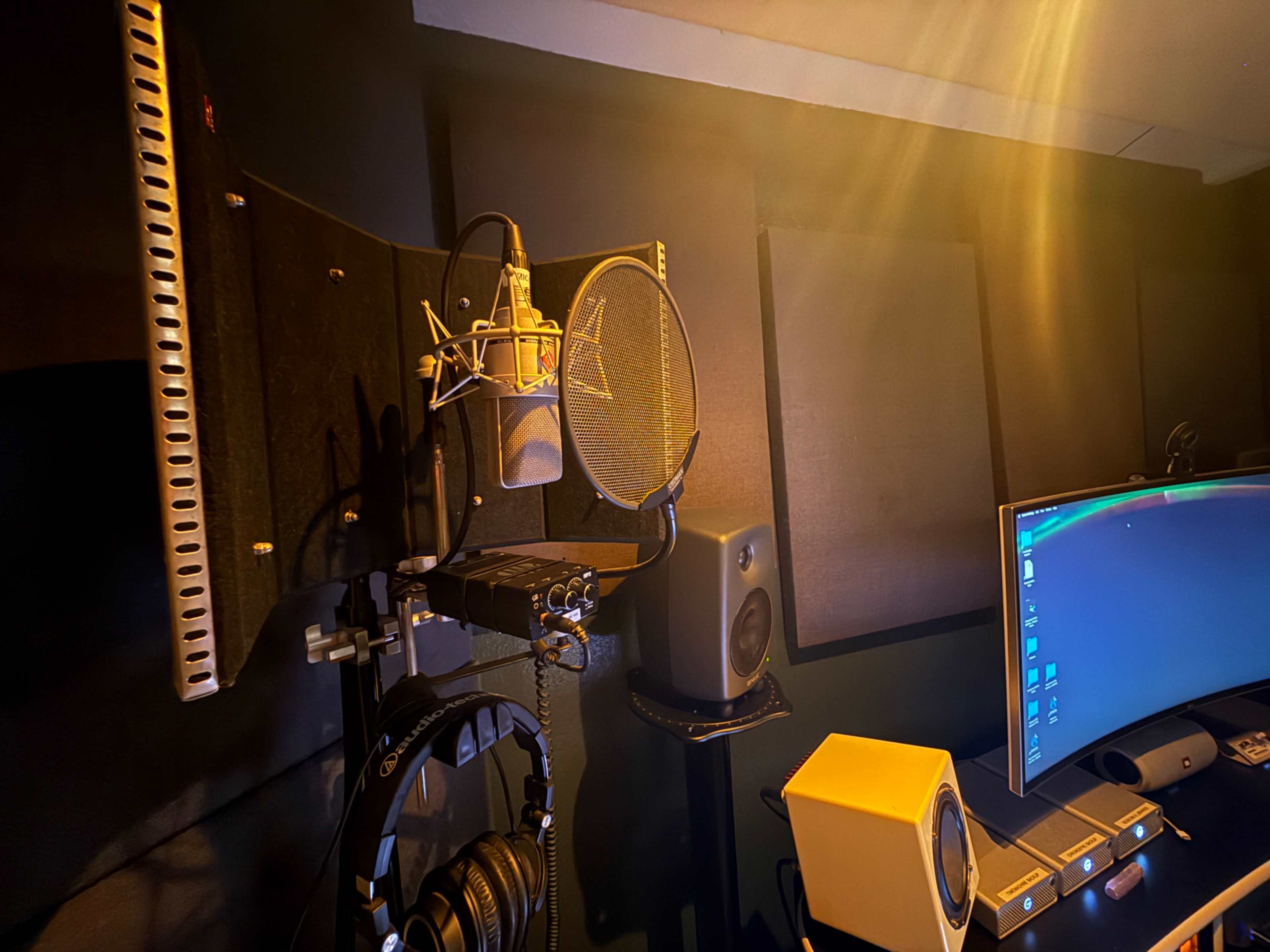 The image shows a recording studio setup featuring a microphone, headphones, soundproof panels, and a computer monitor.