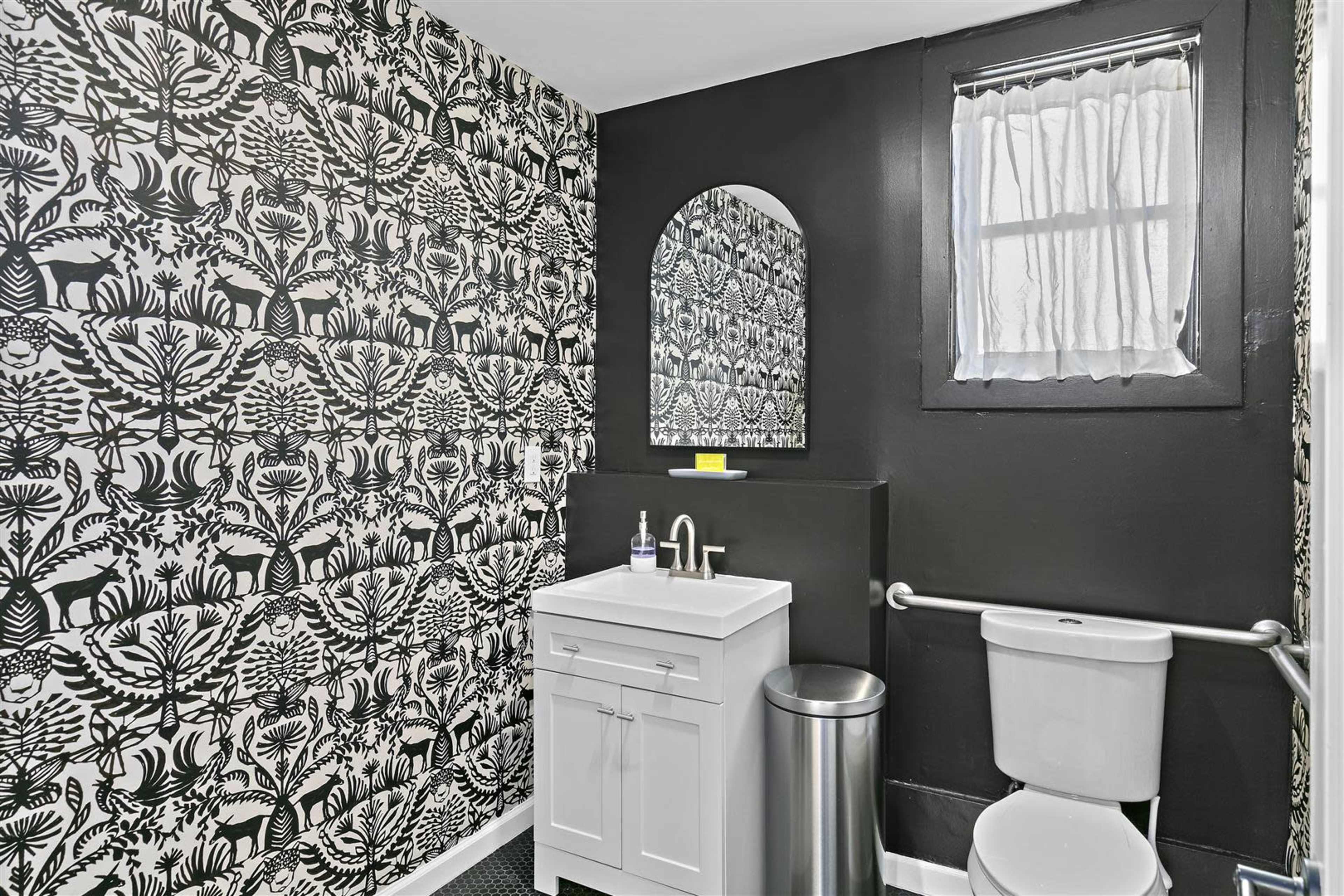 The bathroom features a black and white patterned wallpaper, a modern white vanity with a mirror, and a toilet beside a small window.