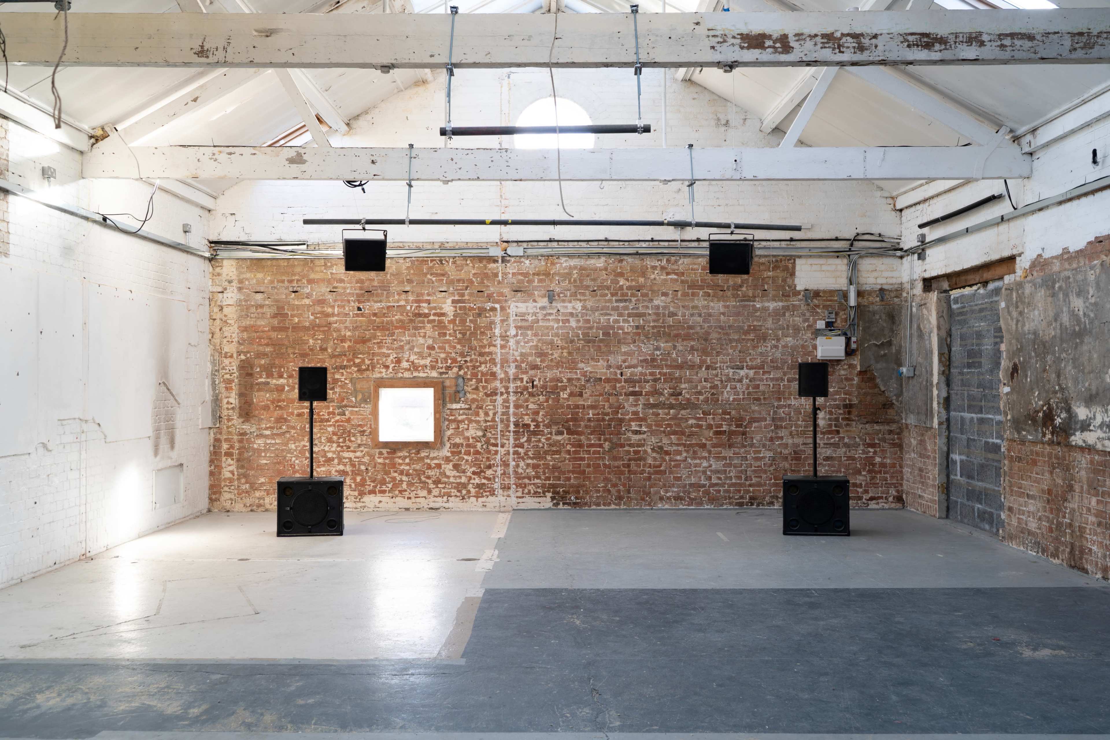 The image shows a spacious interior of a warehouse with exposed brick walls, wooden beams, and four sound speakers positioned in the corner.