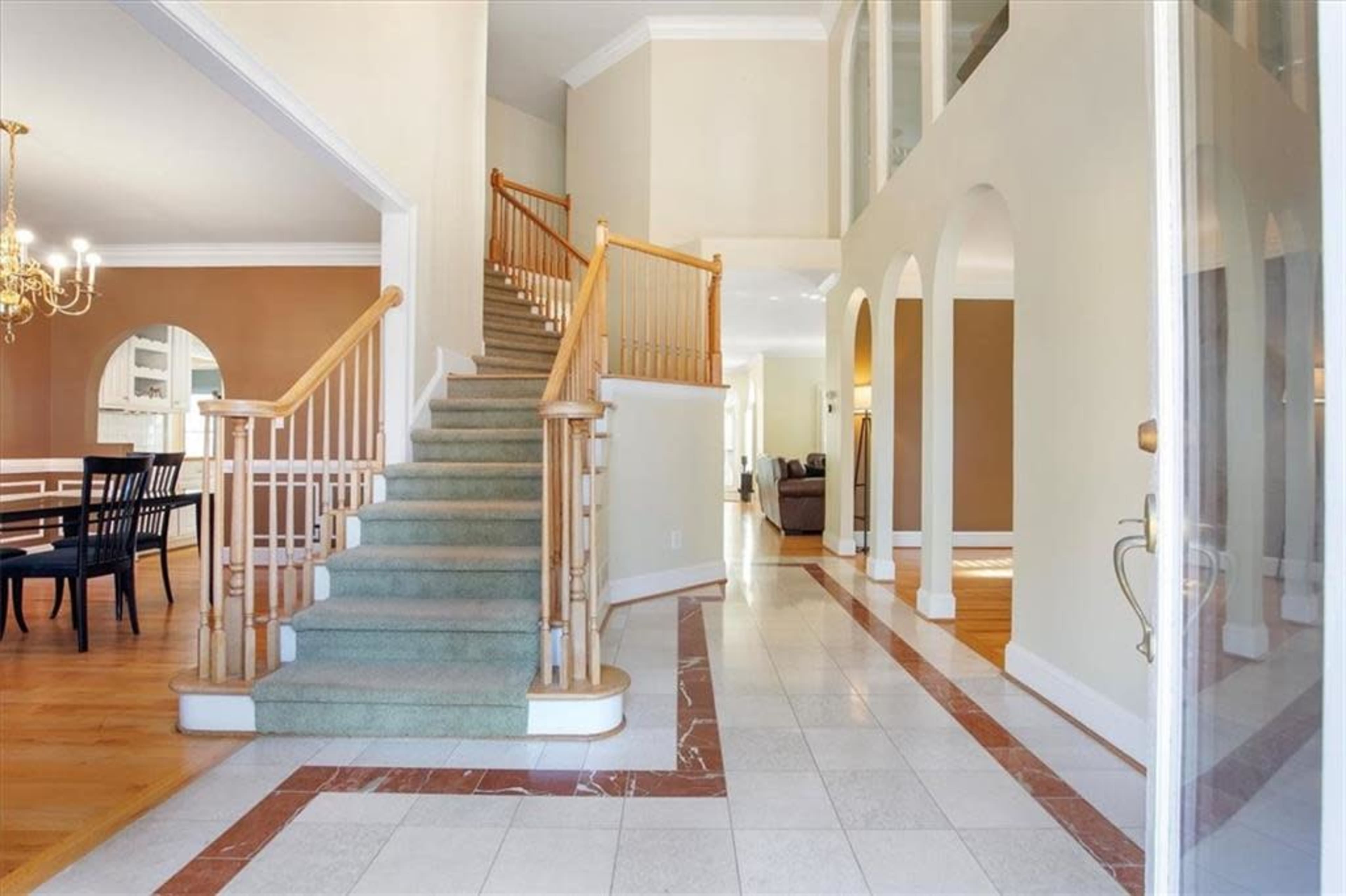 The image shows an entryway featuring a staircase with wooden railings, tiled flooring, and a dining area visible in the background.
