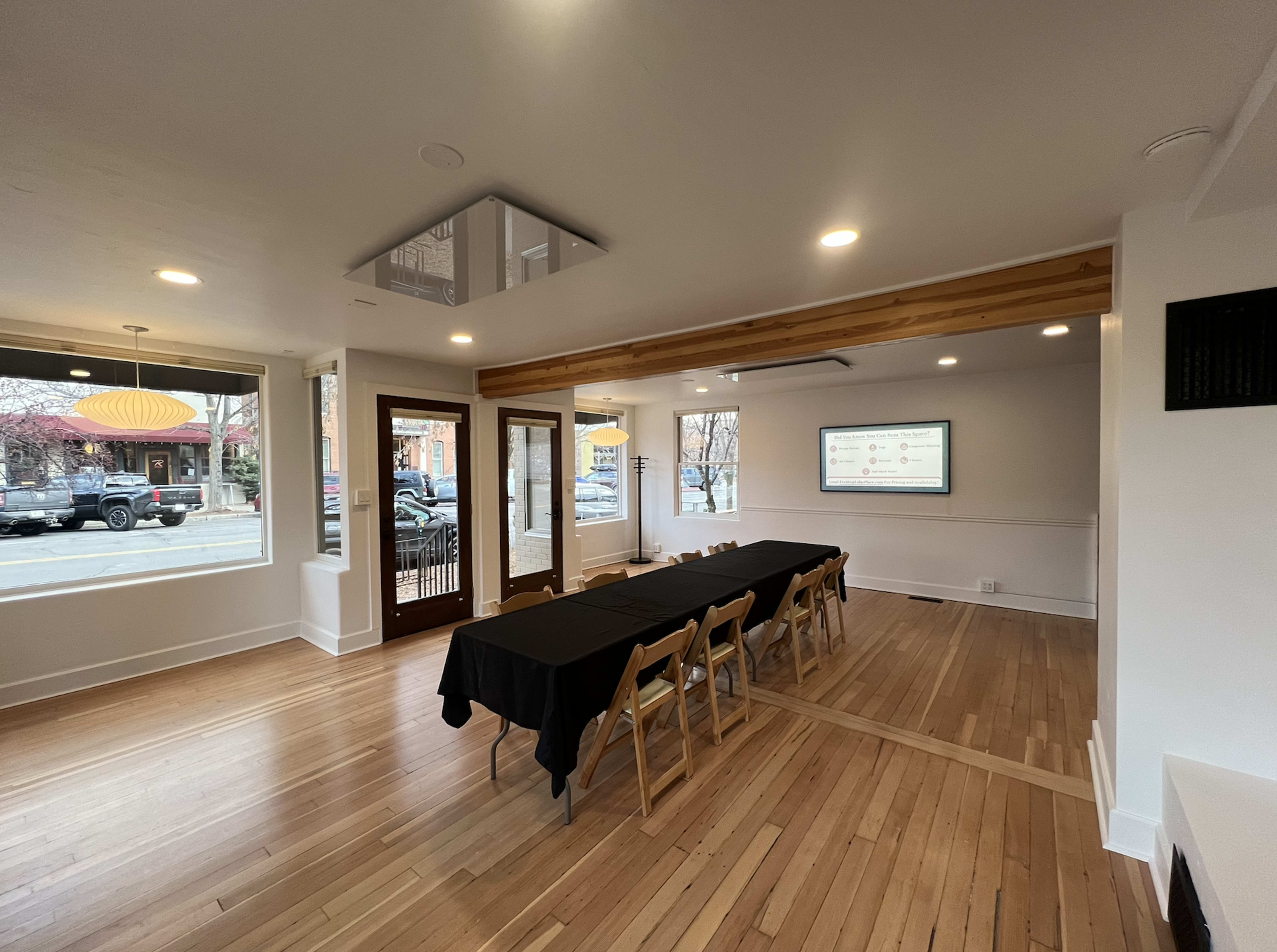 The image shows a spacious, well-lit meeting room with a long black table set up for a presentation, featuring wooden flooring and large windows overlooking the street outside.