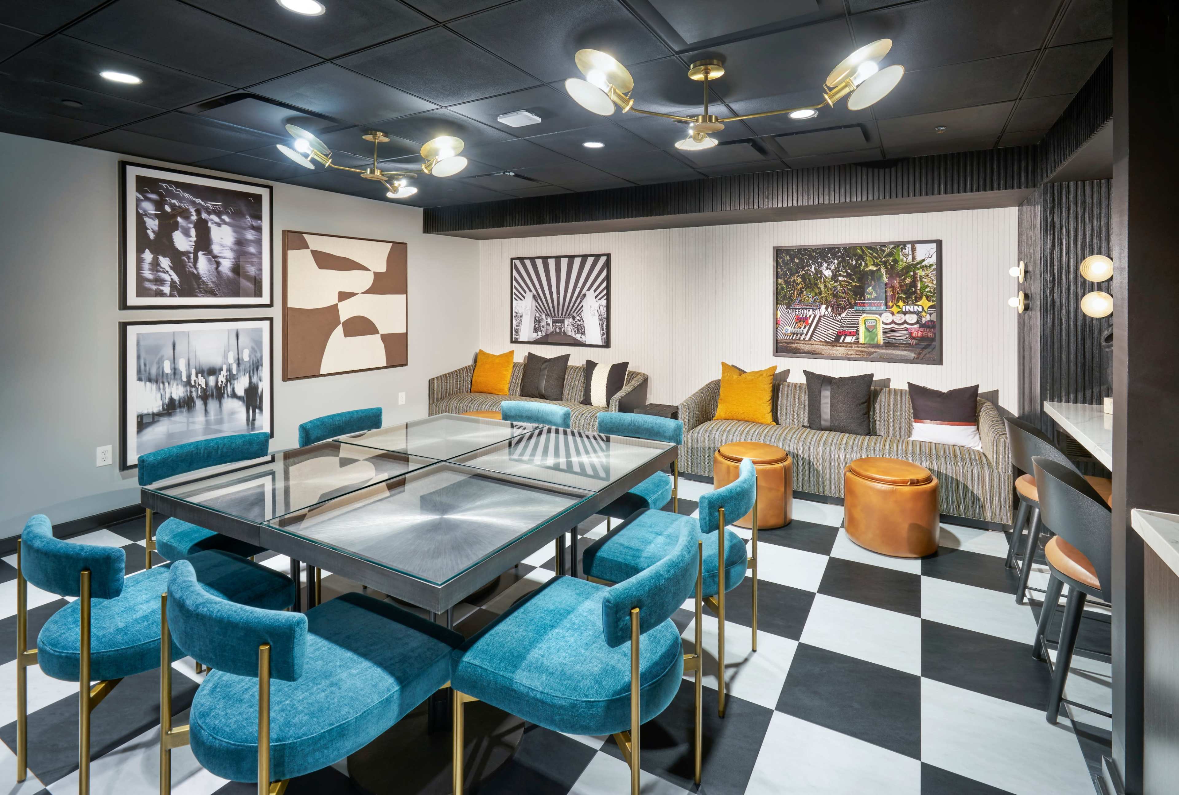 The image shows a contemporary lounge area with a glass table, teal chairs, a sectional couch with decorative pillows, and framed artwork on the walls, all set against a black and white checkered floor.